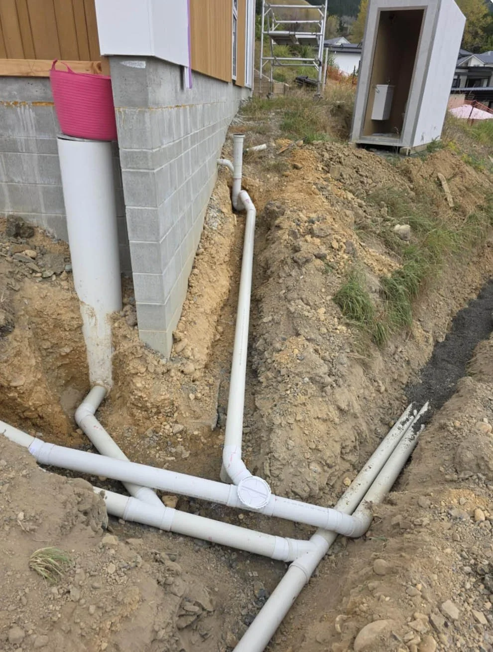 Underground plumbing pipes outside a building under construction, with a concrete block wall, a pink bucket, and a small electrical box in a gray enclosure on a grassy hillside.