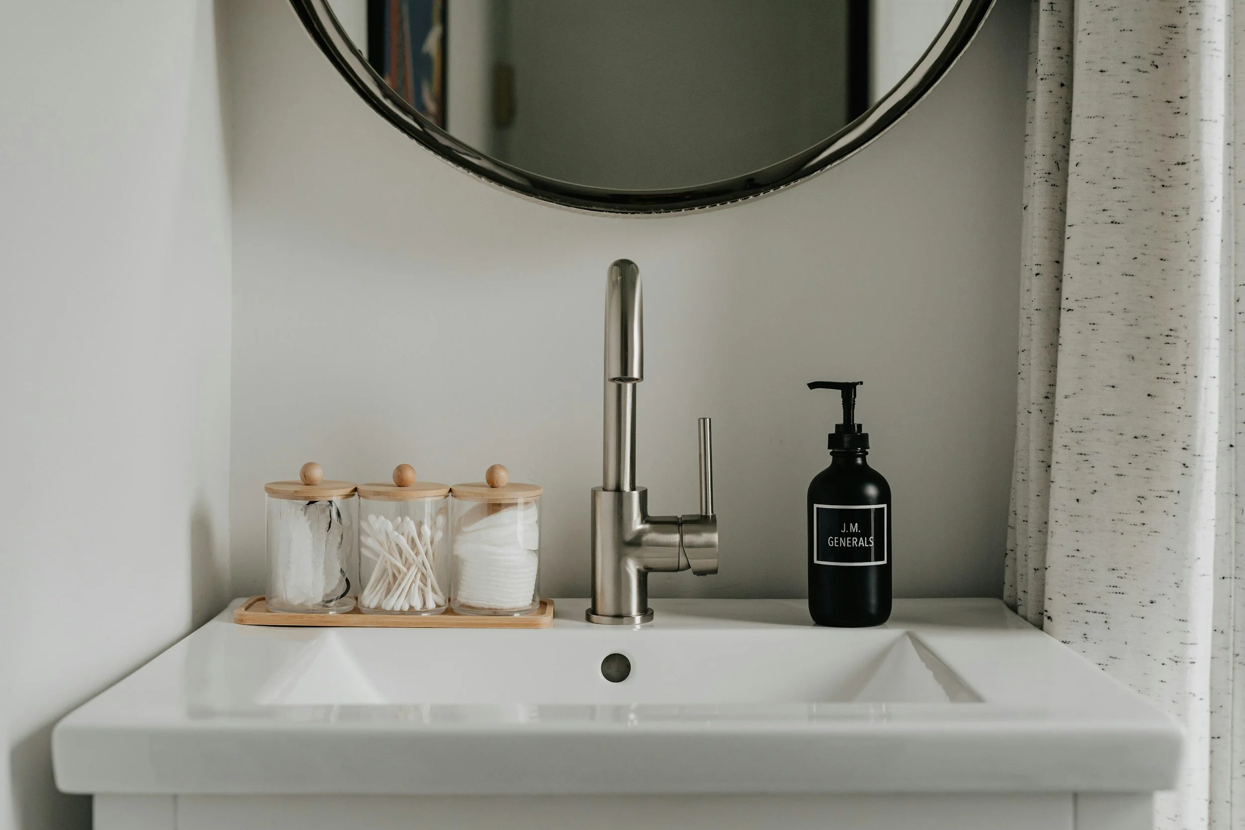 Bathroom sink with a modern chrome faucet, a black soap dispenser labeled 'J.M. Generals', and three glass jars with wooden lids containing cotton swabs, cotton pads, and cotton balls, on a wooden tray, with a round mirror above and white curtains on the side.