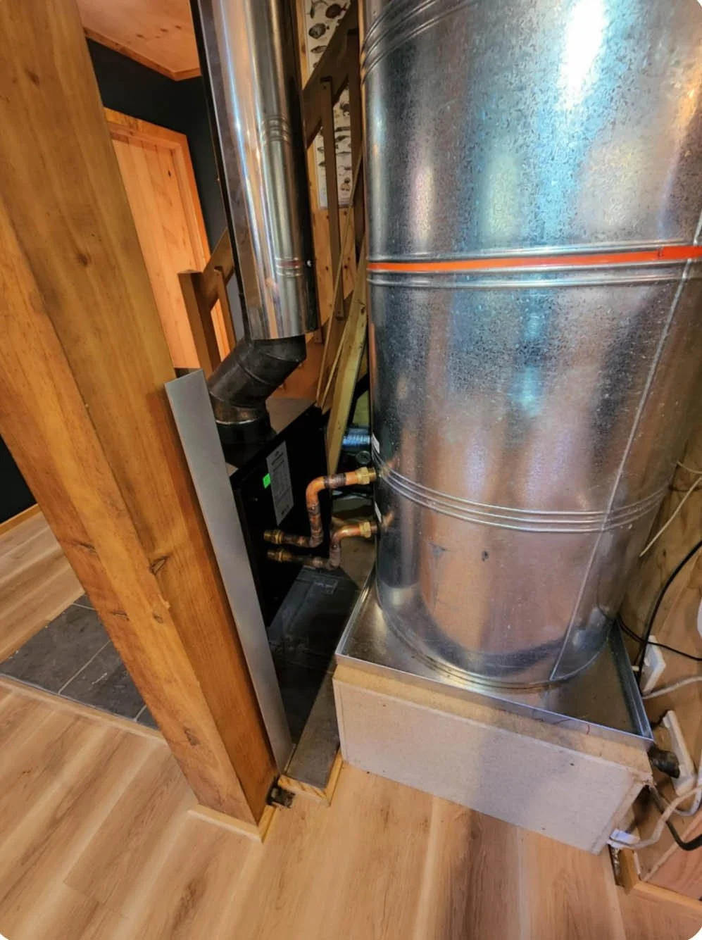 A metal furnace or heater connected to wood framing and copper piping inside a room with wooden floors and walls.