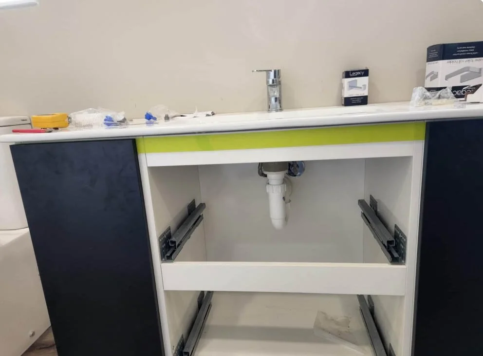 Undermounted kitchen or bathroom sink with a faucet, installed on a countertop with a green stripe, missing cabinet doors, with various small items and packaging on the countertop.