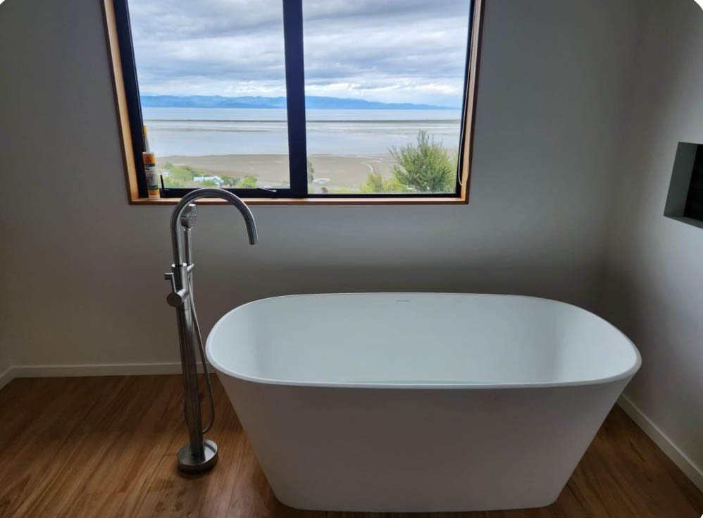 A white freestanding bathtub with a modern chrome faucet, positioned in front of a large window overlooking a body of water and mountains, with a wooden floor and plain white walls.