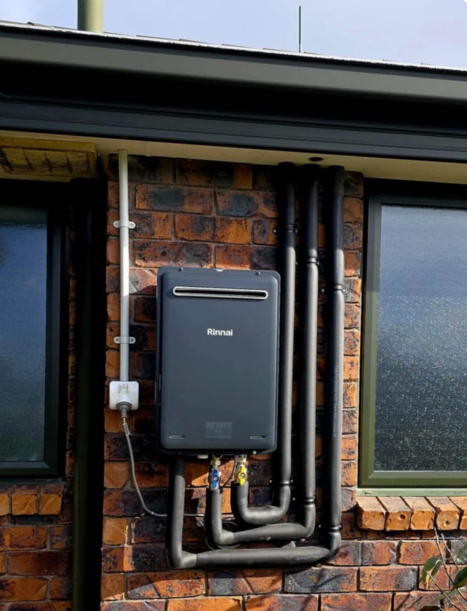 Outdoor Rinnai tankless water heater installed on brick wall with black pipes and electrical wiring.