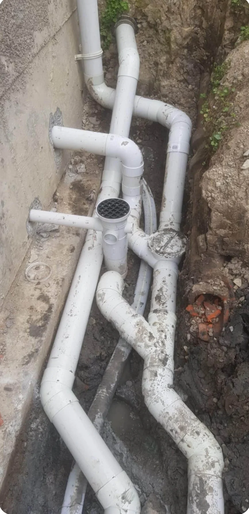 Multiple white PVC pipe connections on a construction site, with some pipes leading into a wall and others extending into the ground.