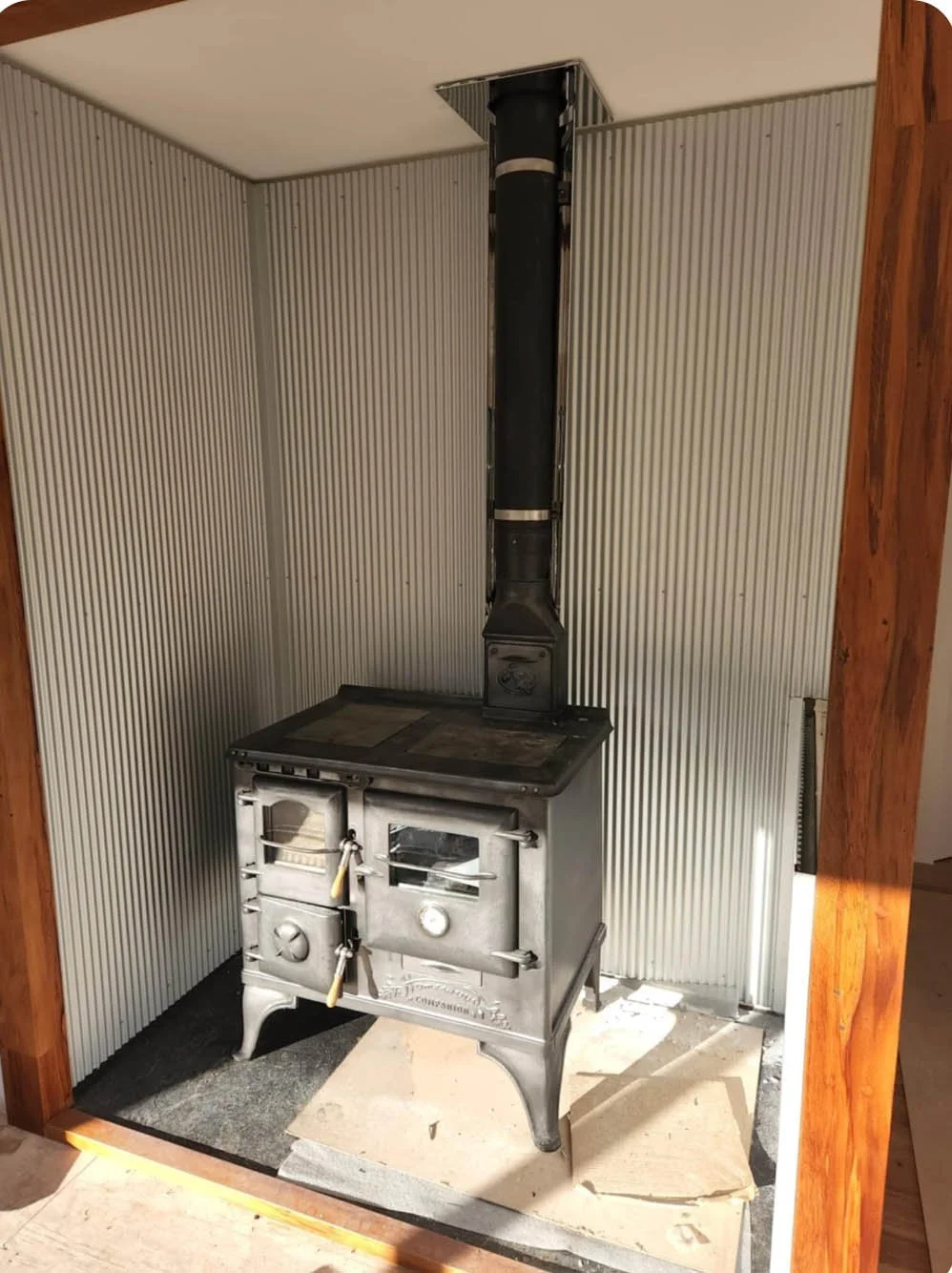 Vintage metal stove with a chimney pipe in a small enclosed space with wood trim and ribbed wall panels.