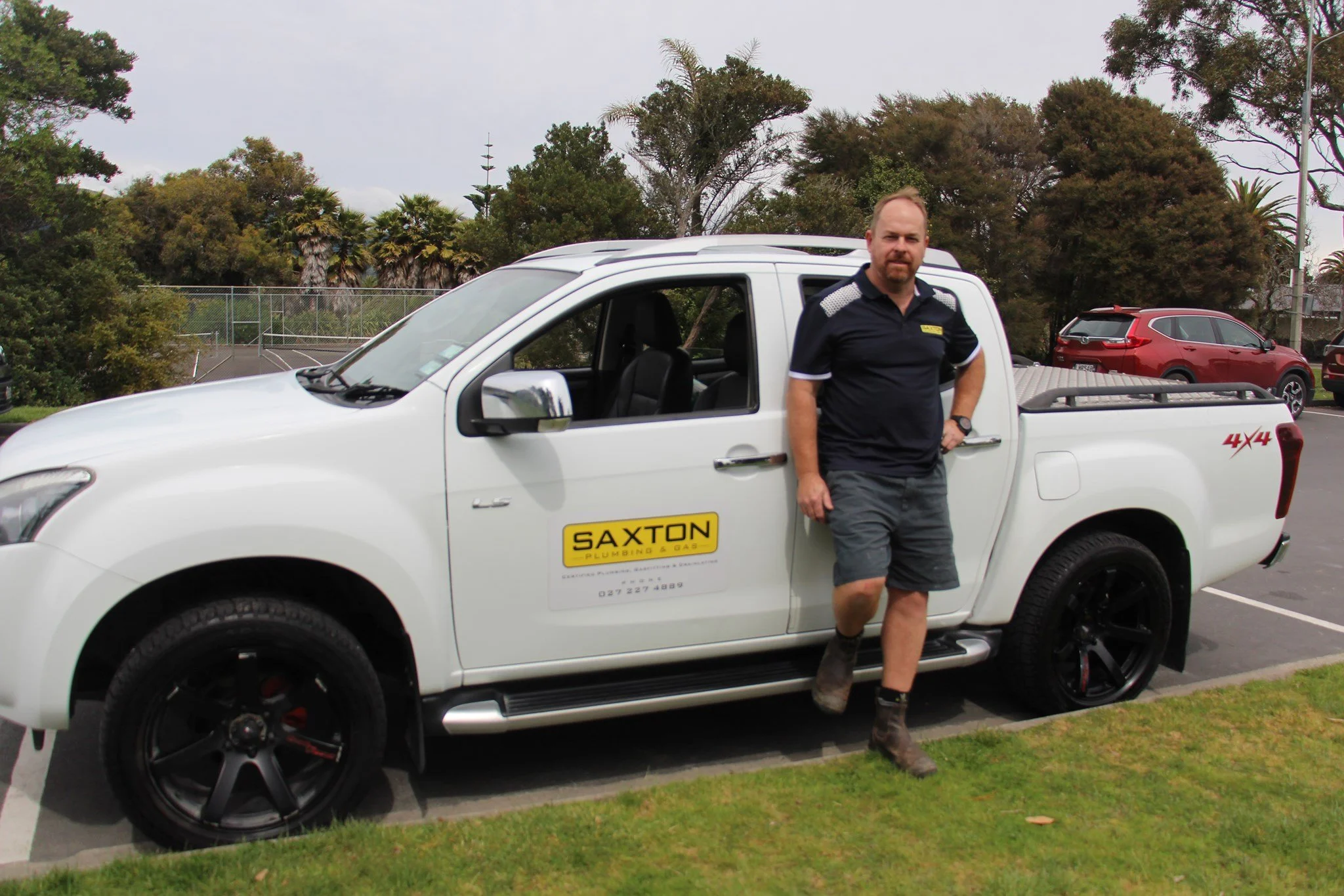 A man standing next to a white pickup truck with a yellow 'Saxton Plumbing & Gas' sign on the door, in a parking lot with trees in the background.