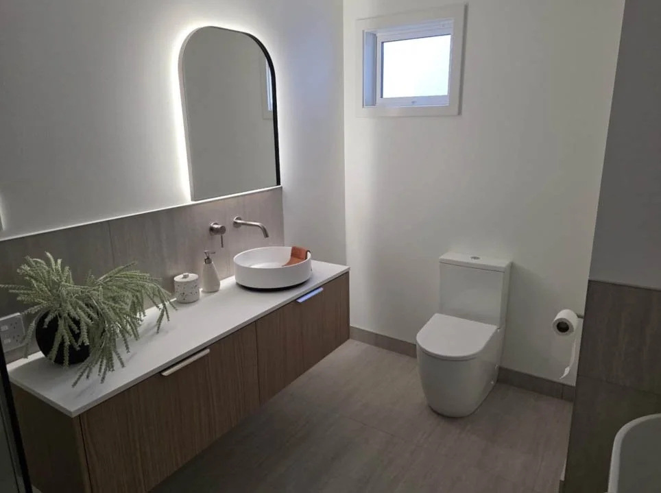 Modern bathroom with a wooden vanity, circular vessel sink, large mirror with backlighting, small window, and a toilet on the right side.