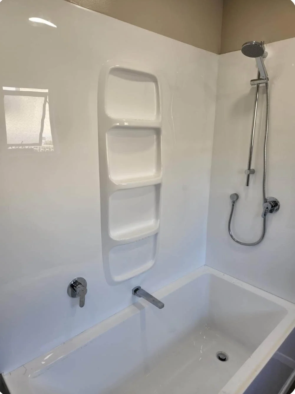 Shower stall with a built-in three-tier white plastic shelf, a handheld shower with a flexible hose, and two knobs likely for water control, with a drain at the bottom.