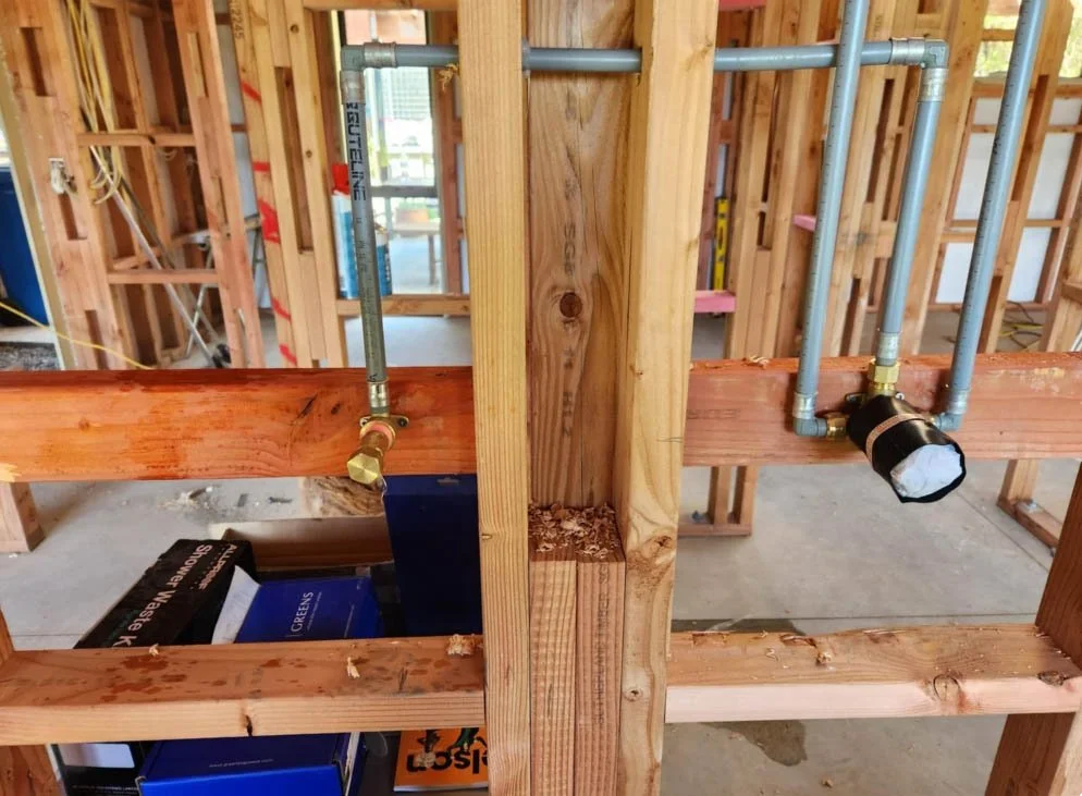 Interior view of a house under construction, showing wooden framing and plumbing pipes installed through the studs.