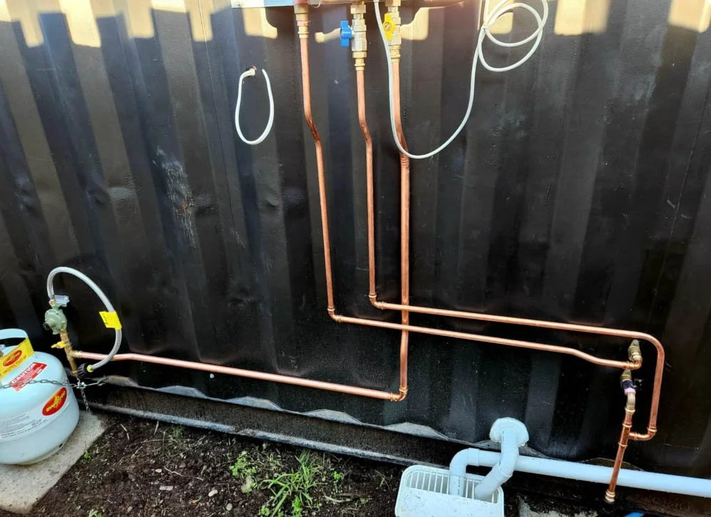 Copper pipes running along a black wall, connected to a gas tank and gray pipe, with some valves and fittings visible.