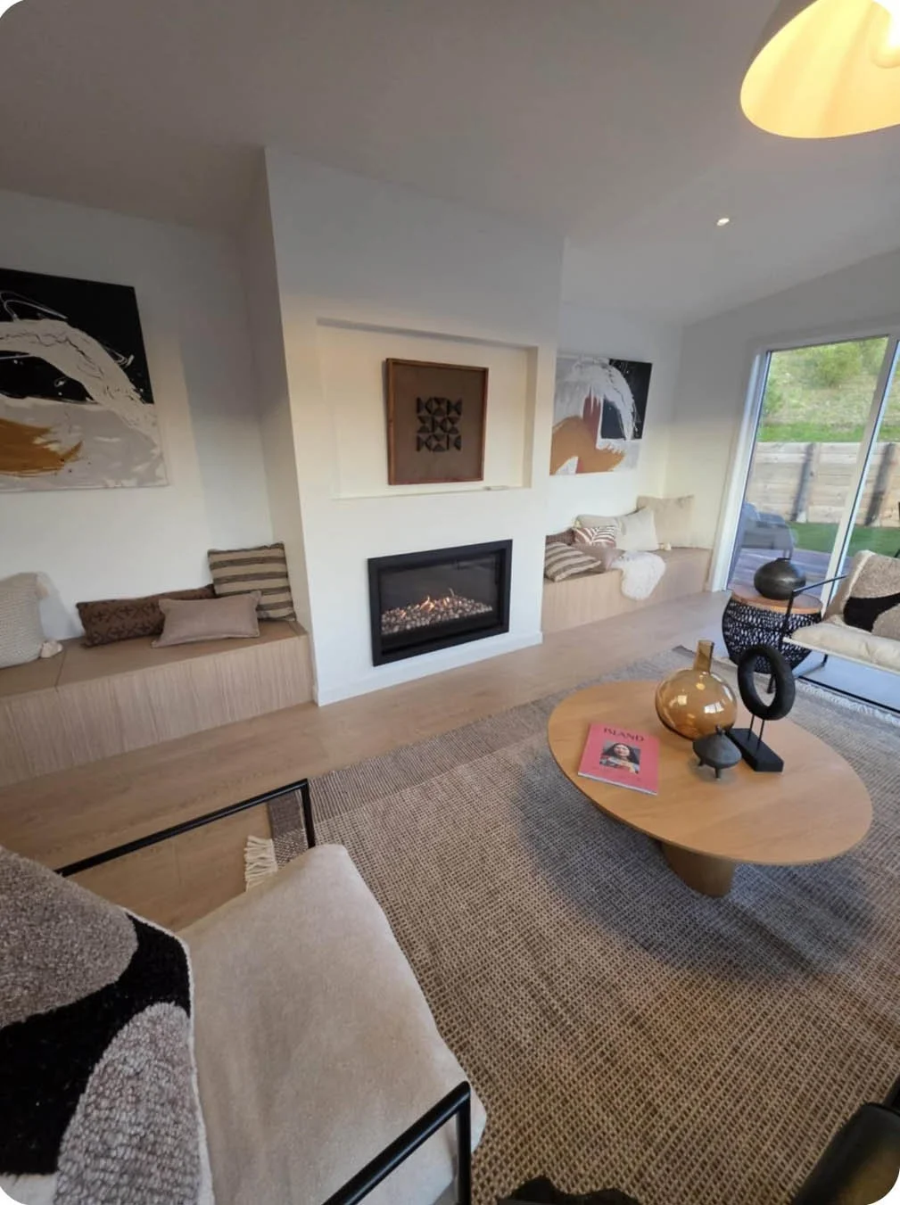 Living room with built-in seating, abstract artwork, a fireplace, a round coffee table with decorative objects, and a sliding glass door leading outside.