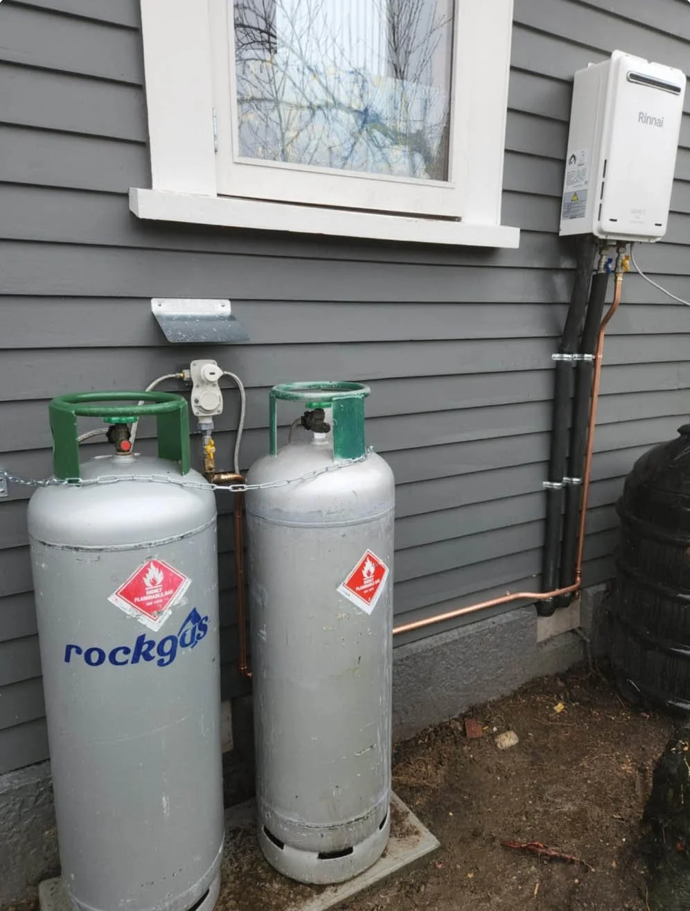 Two large propane tanks with safety labels and gauges next to a gray house wall, with copper pipes connected to a Rinnai tankless water heater mounted on the wall.