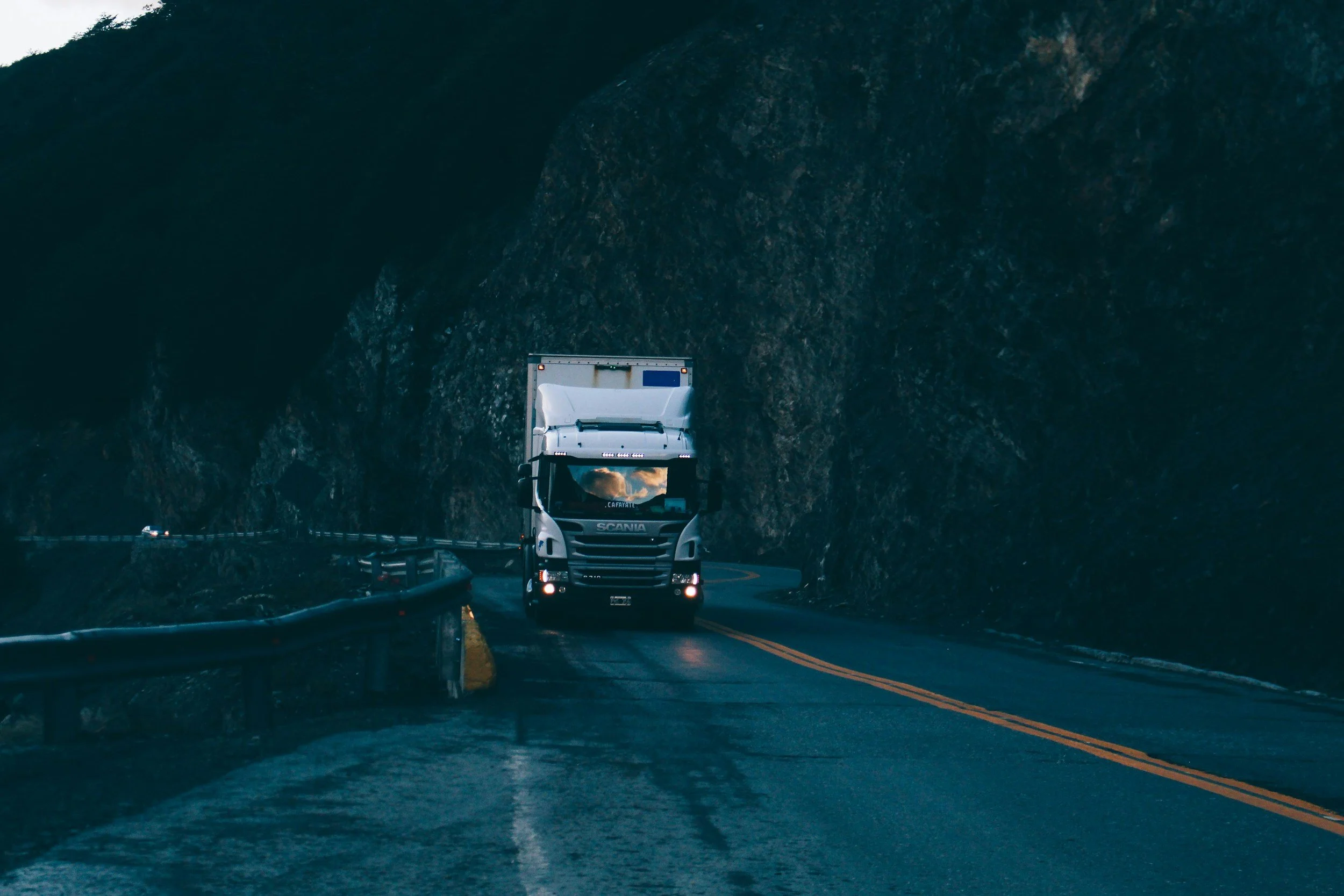 A white semi-truck driving along a winding mountain road with a rocky hillside on one side.