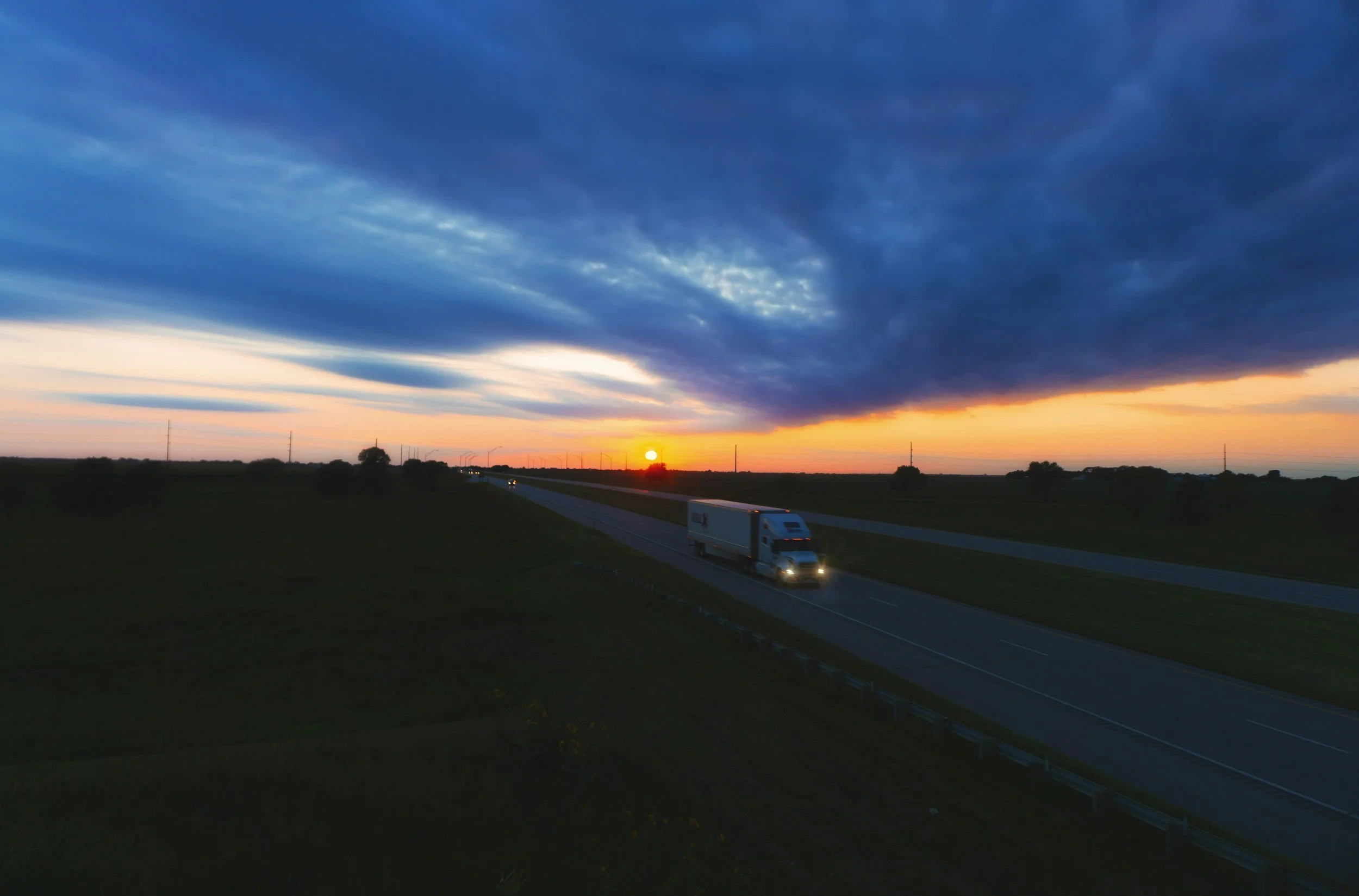 A highway at sunset with a truck traveling on the road, under a sky filled with dark blue and orange clouds.