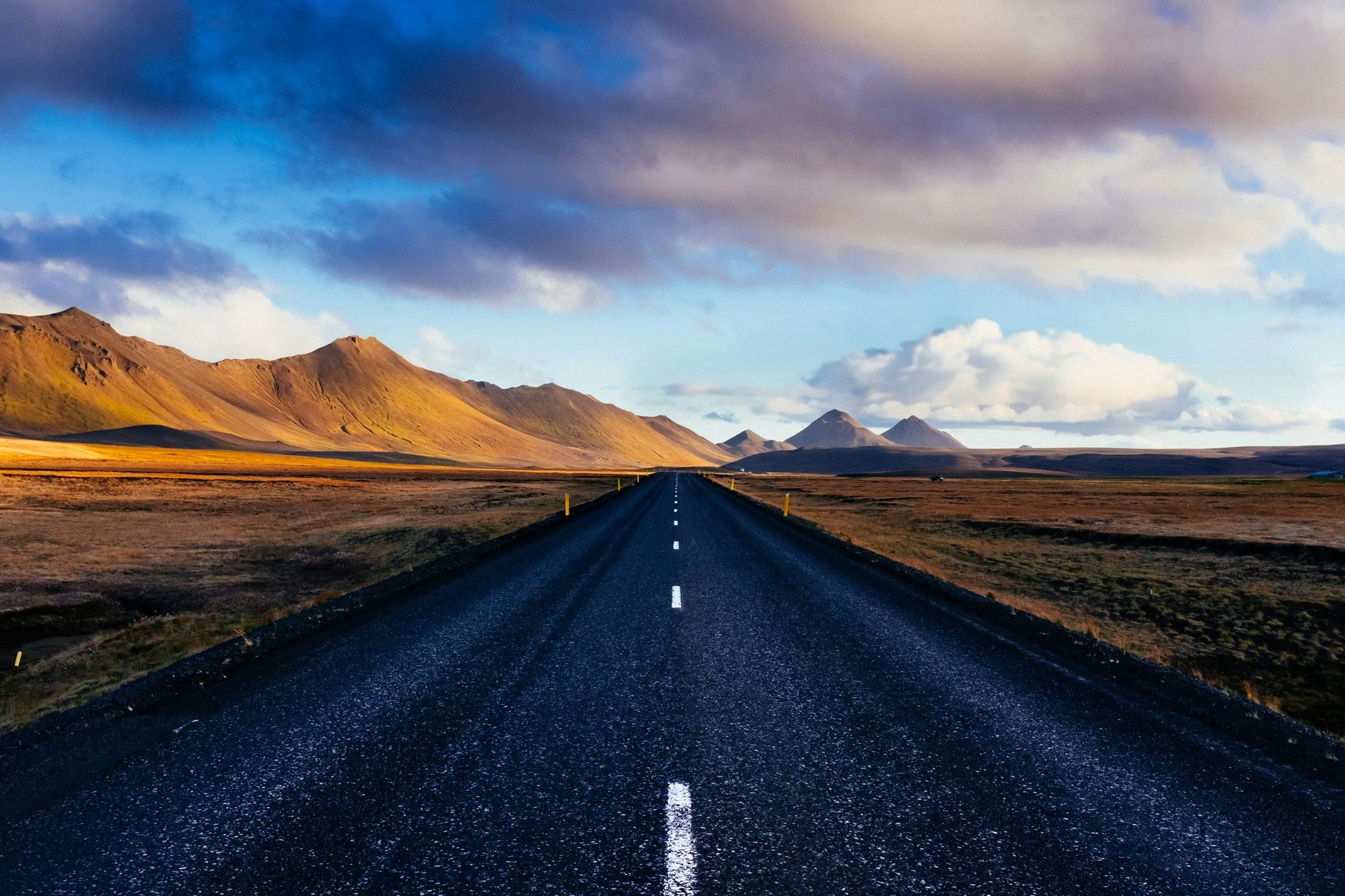 A straight asphalt road extending into the distance with mountains on either side and a cloudy sky overhead.