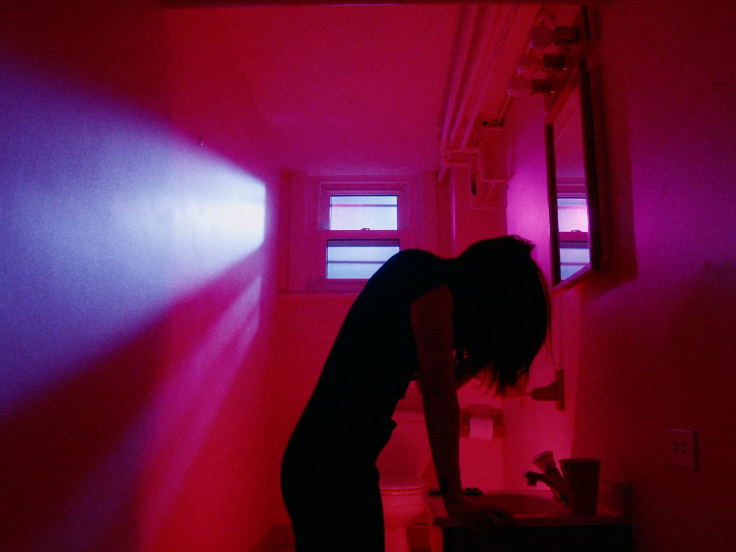 Person with shoulder-length hair leaning over a sink in a dimly lit room with pink and purple lighting, a small window, and a mirror on the wall.