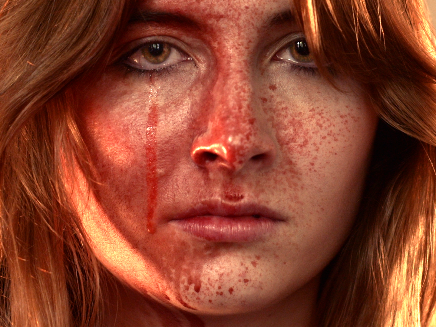 Close-up of a woman's face with red freckles, brown eyes, and long red hair, with blood streaks on her cheek and nose.
