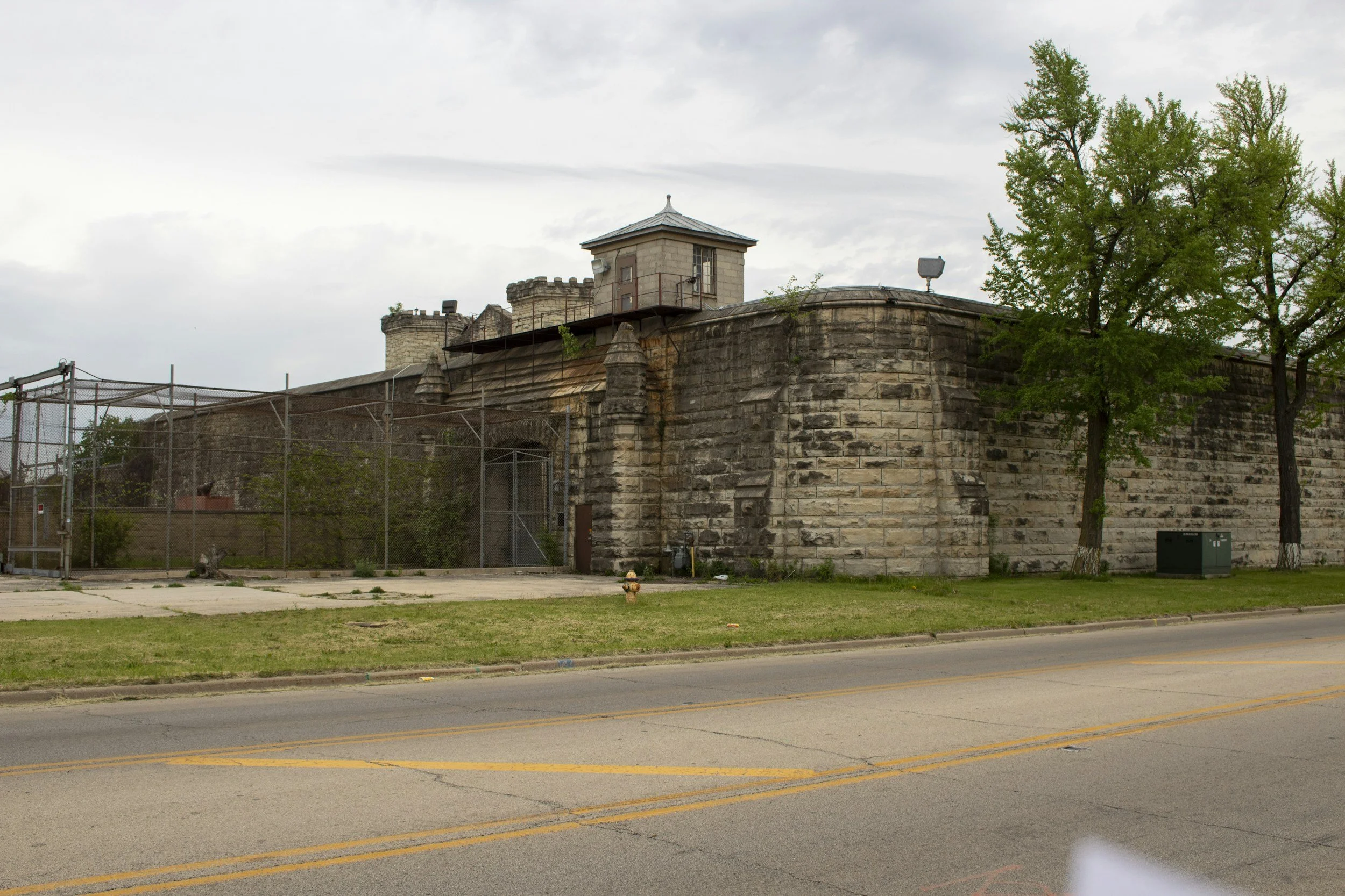A large stone fortress wall with a small tower on top, trees on the right side, a grassy area in front, and a tennis court to the left, under an overcast sky.