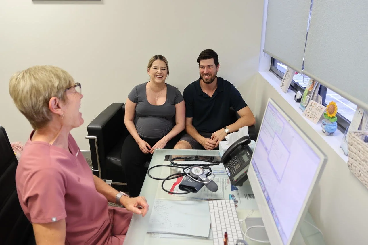 A doctor, two patients, a pregnant woman and a man, sitting in an office with a desk, computer, phone, and medical equipment, having a conversation and smiling.