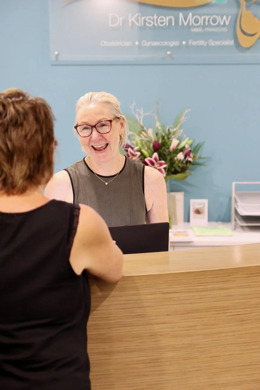 A woman with glasses and blonde hair smiling at a receptionist behind a wooden counter. The receptionist has short, light-colored hair and is wearing a sleeveless top. Behind her is a blue wall with a sign that reads 'Dr Kirsten Morrow' and a colorful bouquet of flowers on the counter.