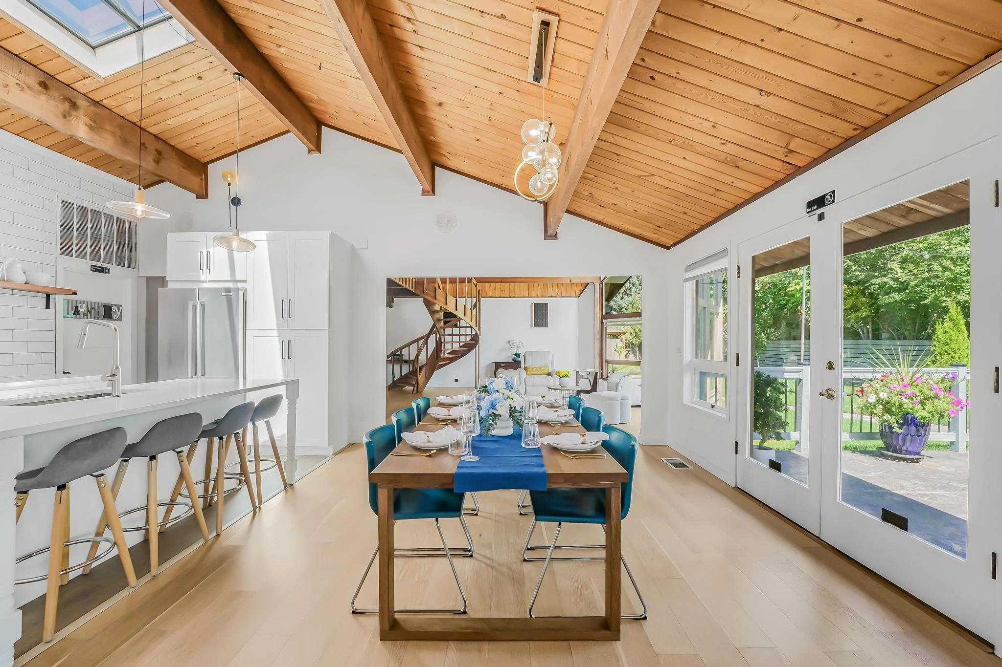 Open-plan dining and kitchen area with wooden vaulted ceiling, large windows showing a backyard with flowering plants, a dining table set for a meal, and a spiral staircase leading to an upper level.