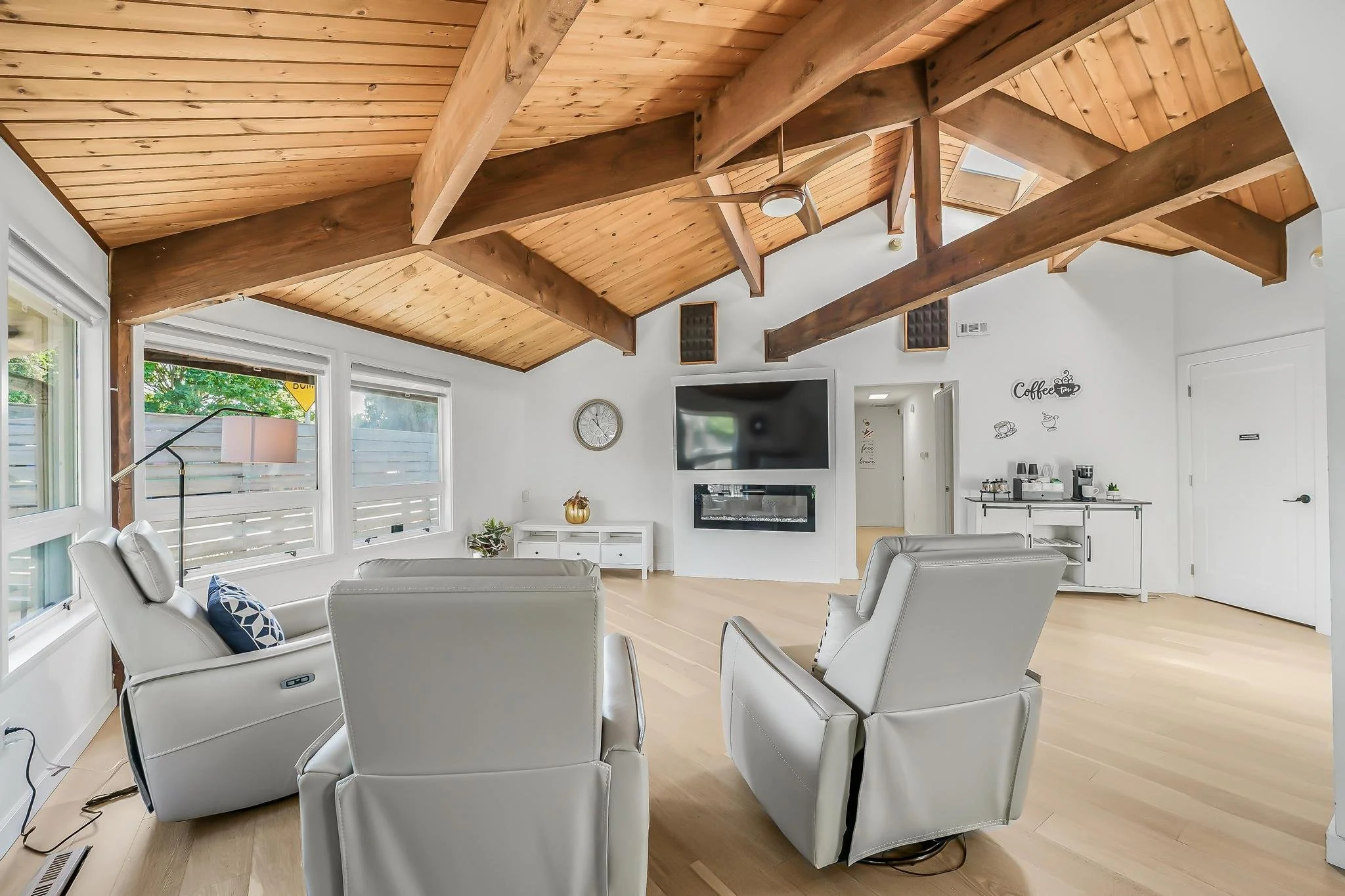 Living room with white walls, three white recliner chairs, wooden vaulted ceiling, and large windows with blinds. A flat-screen TV is mounted on the wall, and a modern fireplace is below it. There is a white console table, a small coffee station, and a clock on the wall.