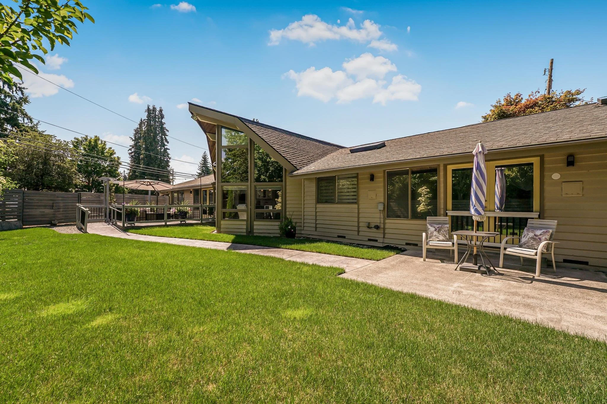 Backyard with green lawn, concrete patio, outdoor furniture with striped umbrellas, and house with large windows and a sloped roof under a partly cloudy sky.