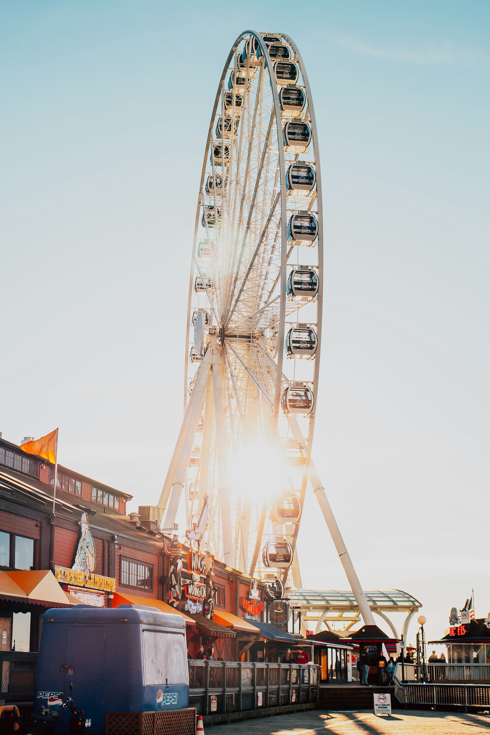 Photo of a large Ferris wheel at a pier or amusement park during sunset, with a building and people below.