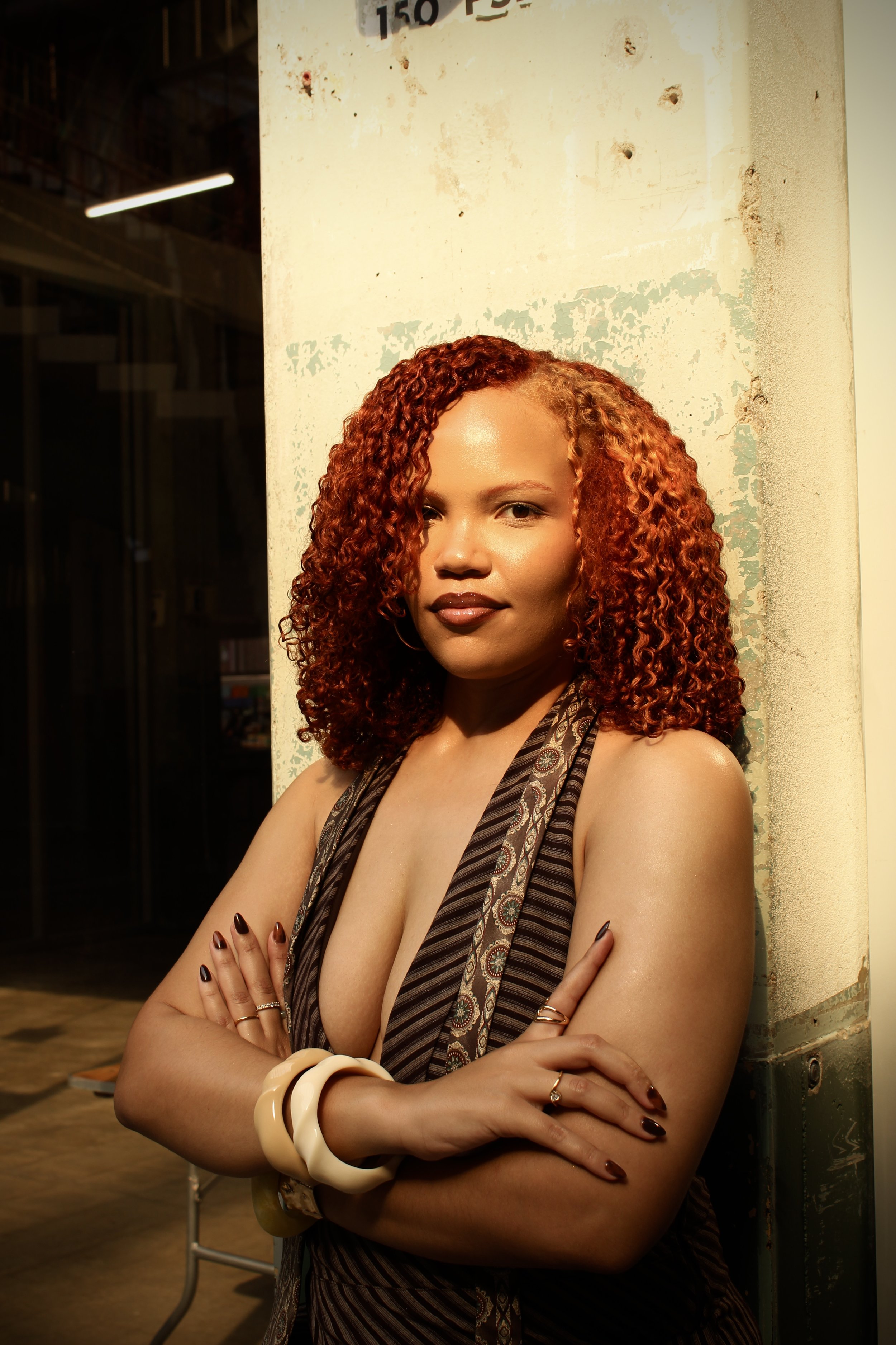 A woman with curly red hair leaning against a concrete wall with peeling paint, her arms crossed, wearing a sleeveless dress with a patterned scarf, dark lipstick, and multiple rings and bracelets, in an industrial setting.
