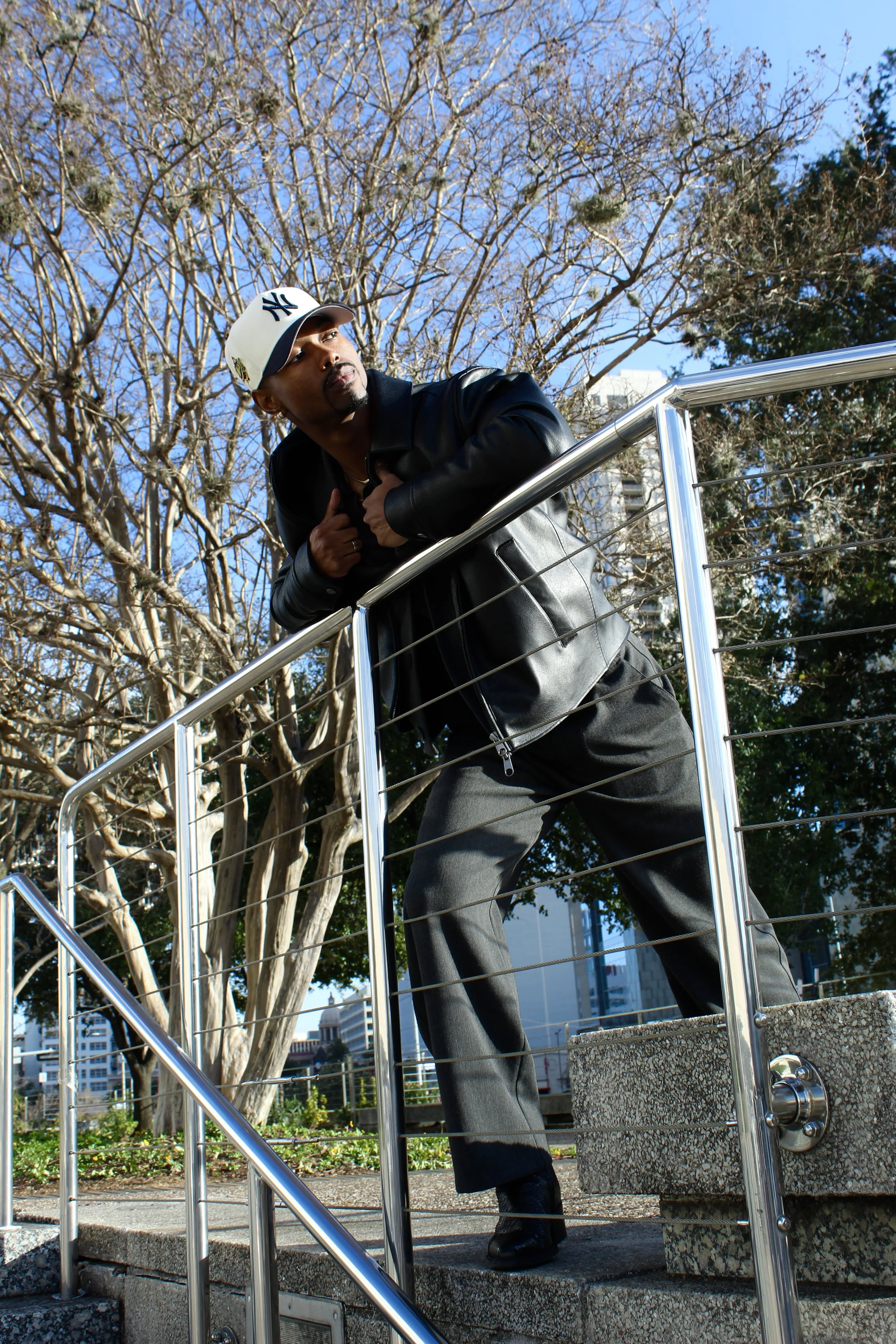 A man wearing a white New York Yankees baseball cap, black leather jacket, and dark pants leaning on a metal railing outdoors during daytime. Trees and buildings are visible in the background, with blue sky overhead.