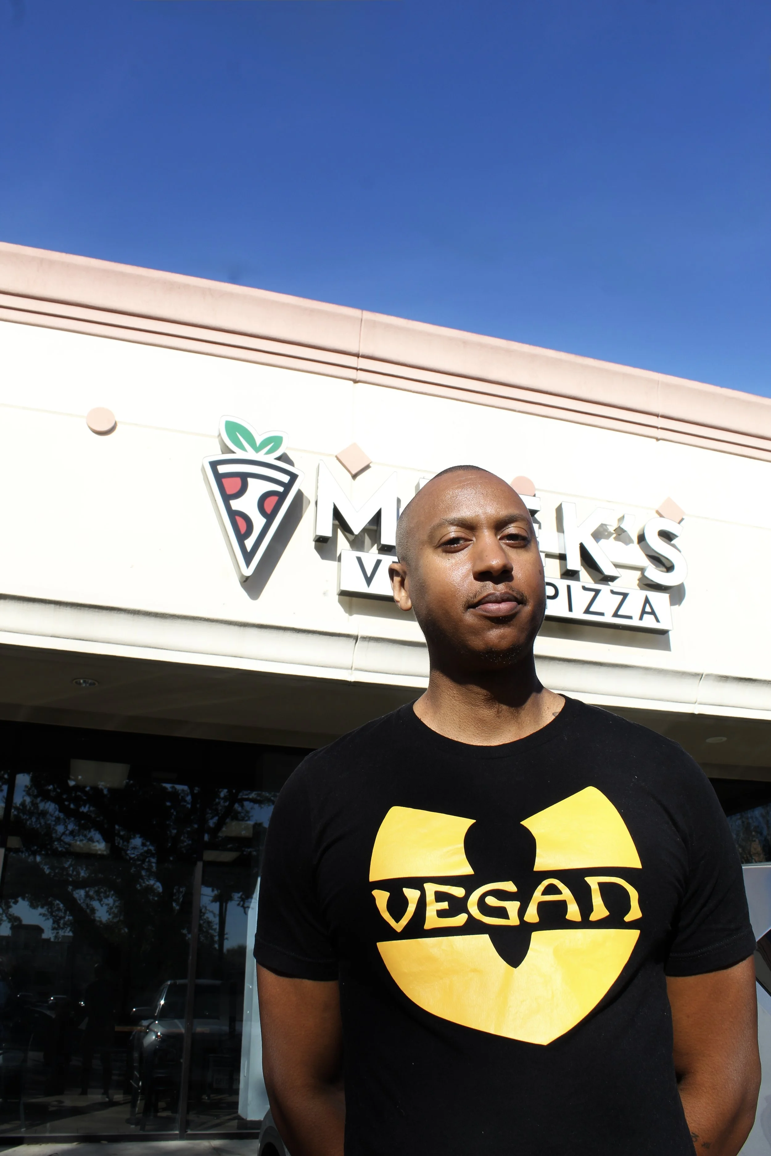 A person stands outside a pizza restaurant on a sunny day. The person is wearing a black t-shirt with a yellow vegan symbol resembling a superhero emblem. The restaurant sign behind them displays a pizza slice icon and the words 'MUNCHIES PIZZA'.