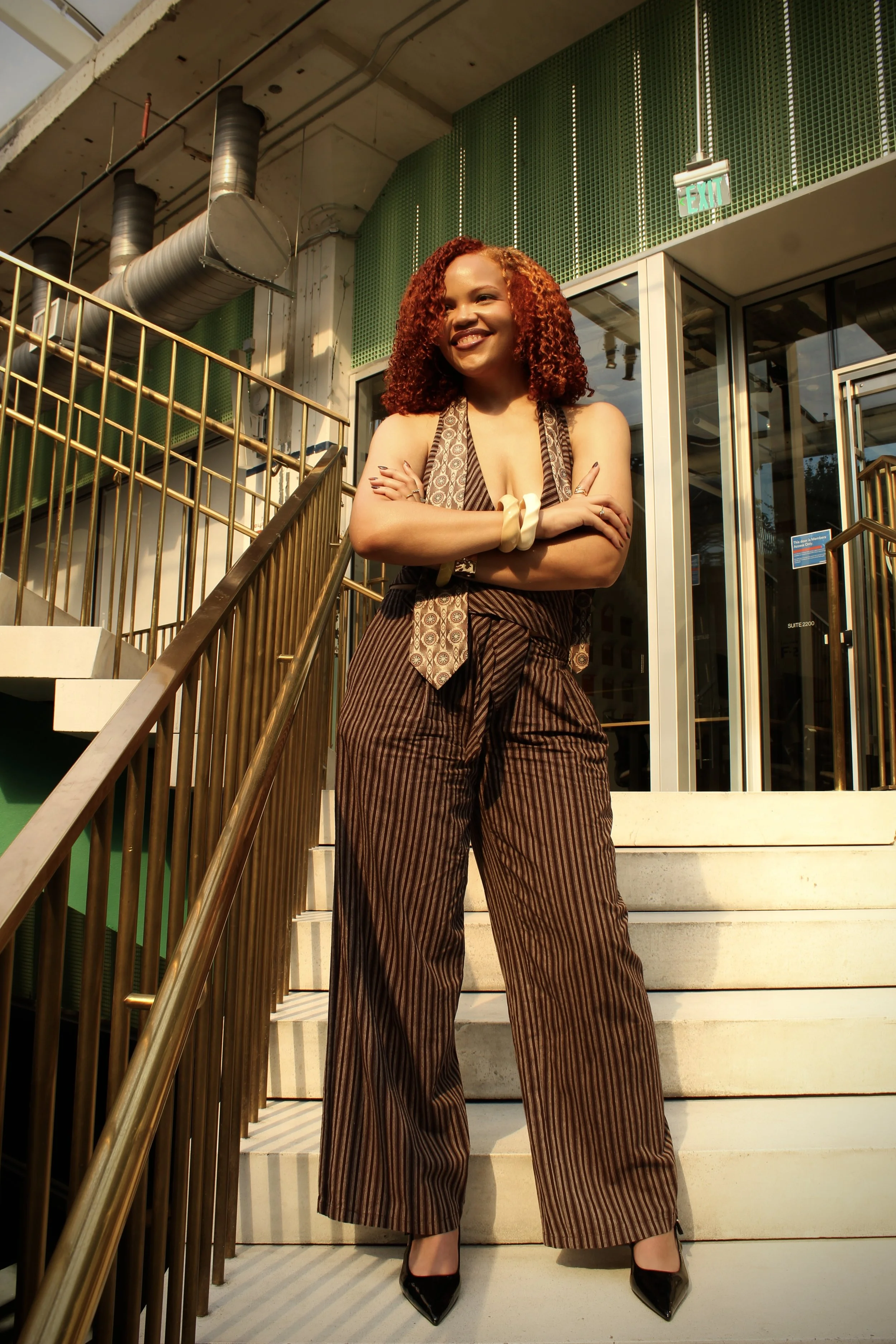 A woman with curly red hair standing on stairs outside a building with glass doors, smiling with her arms crossed. She is wearing brown striped wide-leg pants, a patterned scarf, and black high heels.
