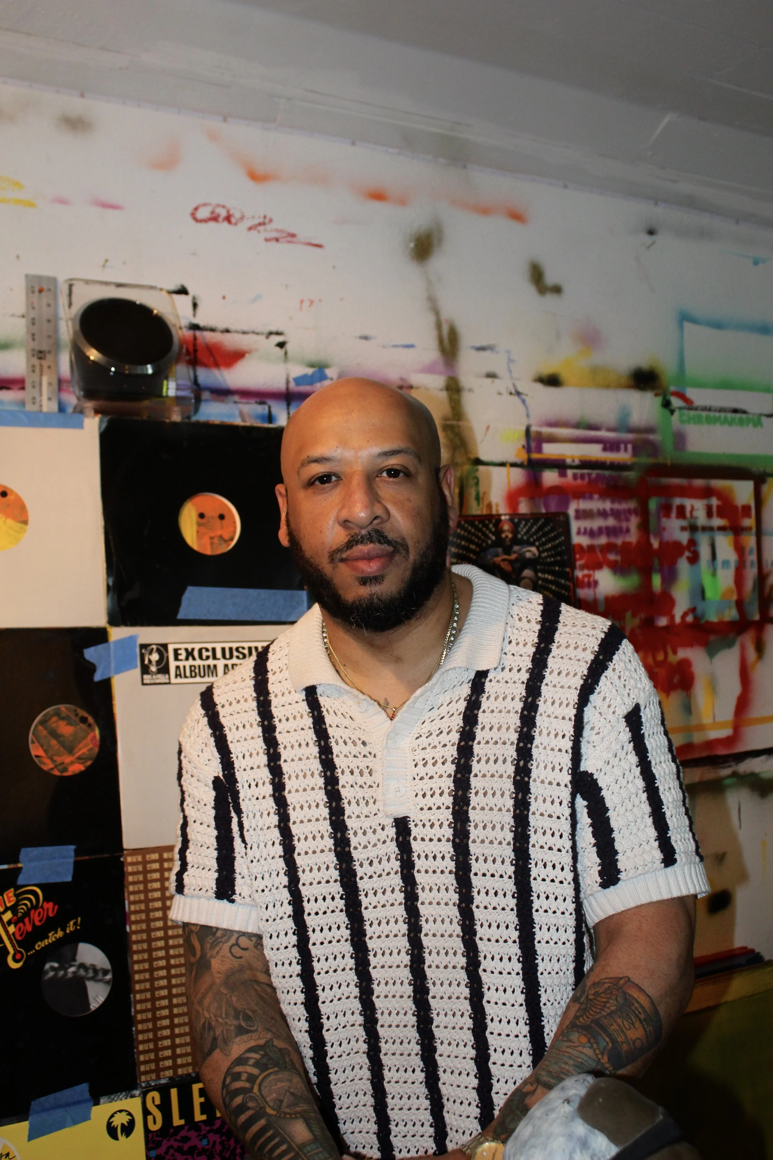 A man with a tattooed arm, light brown complexion, and a beard, standing in front of a colorful, graffiti-like background. He is wearing a white, knitted, black-stripe patterned short-sleeve shirt and gold jewelry.
