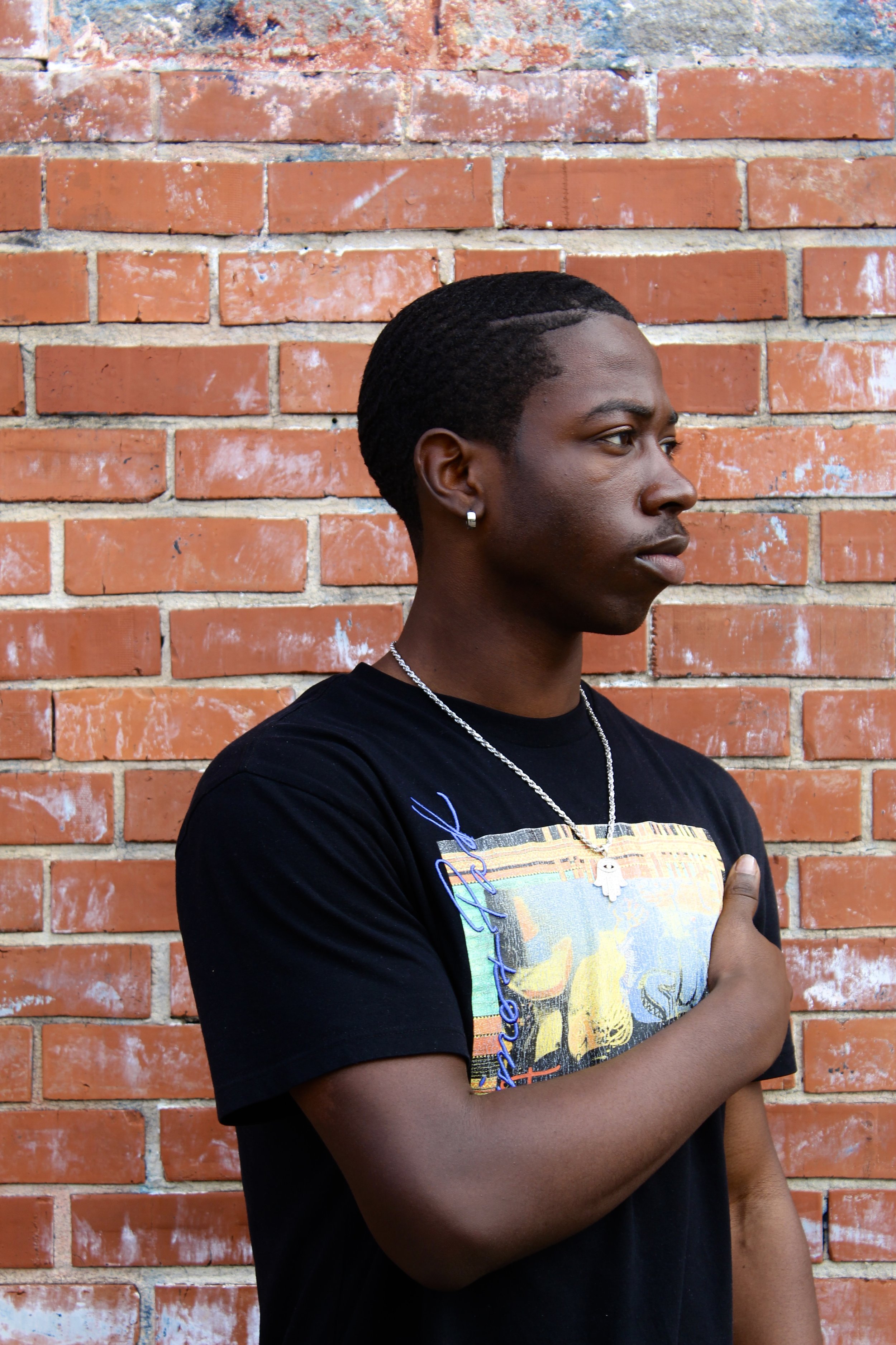 A young man with dark skin and short wavy hair, wearing a black T-shirt with a colorful graphic print, silver jewelry including earrings and a necklace with a pendant, standing against a red brick wall with some white paint splatters, with his arms crossed and looking to the right.