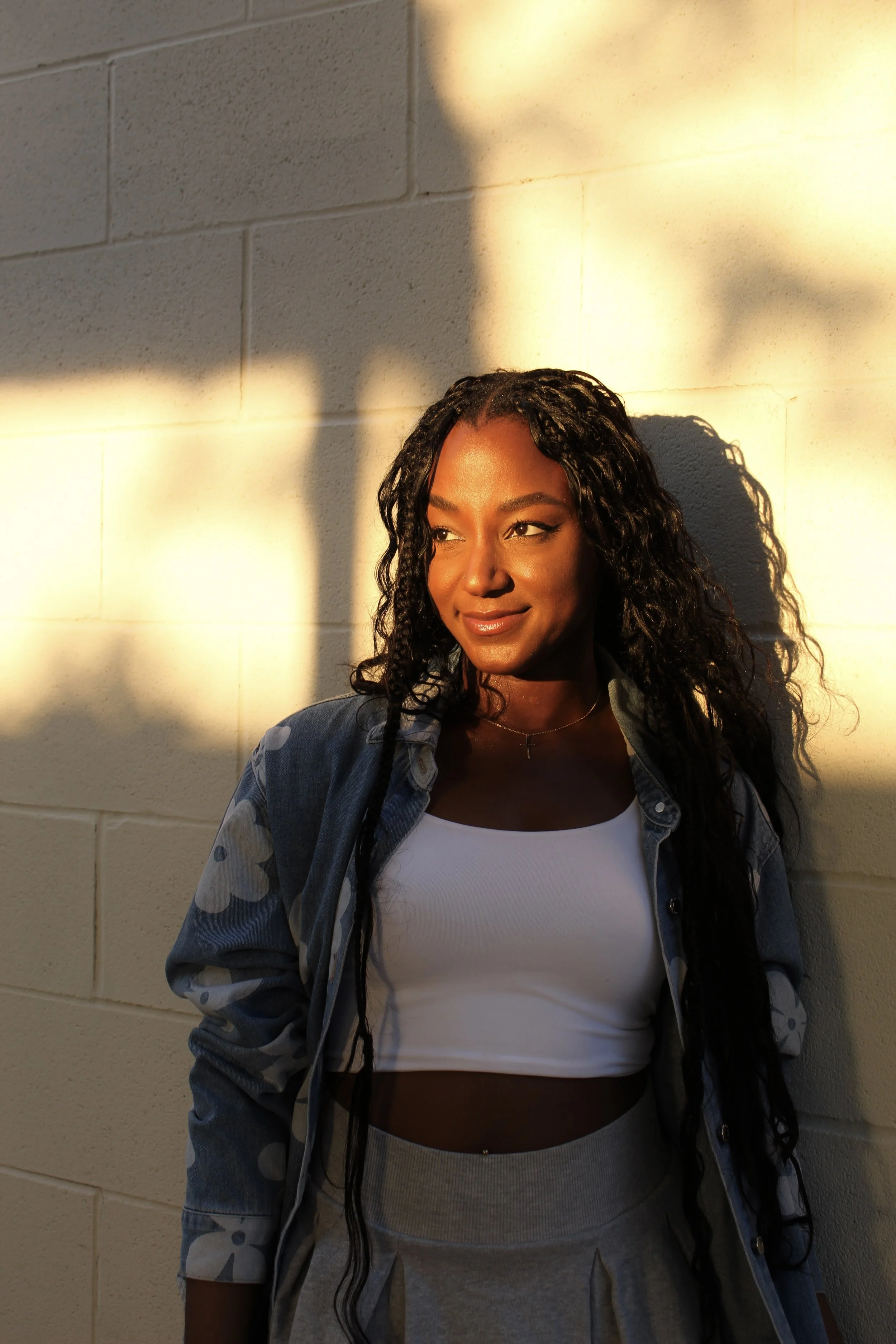 A woman with long curly hair, wearing a white crop top, gray sweatpants, and a blue jacket with a floral pattern, standing against a beige brick wall with sunlight casting shadows on her face and the wall.