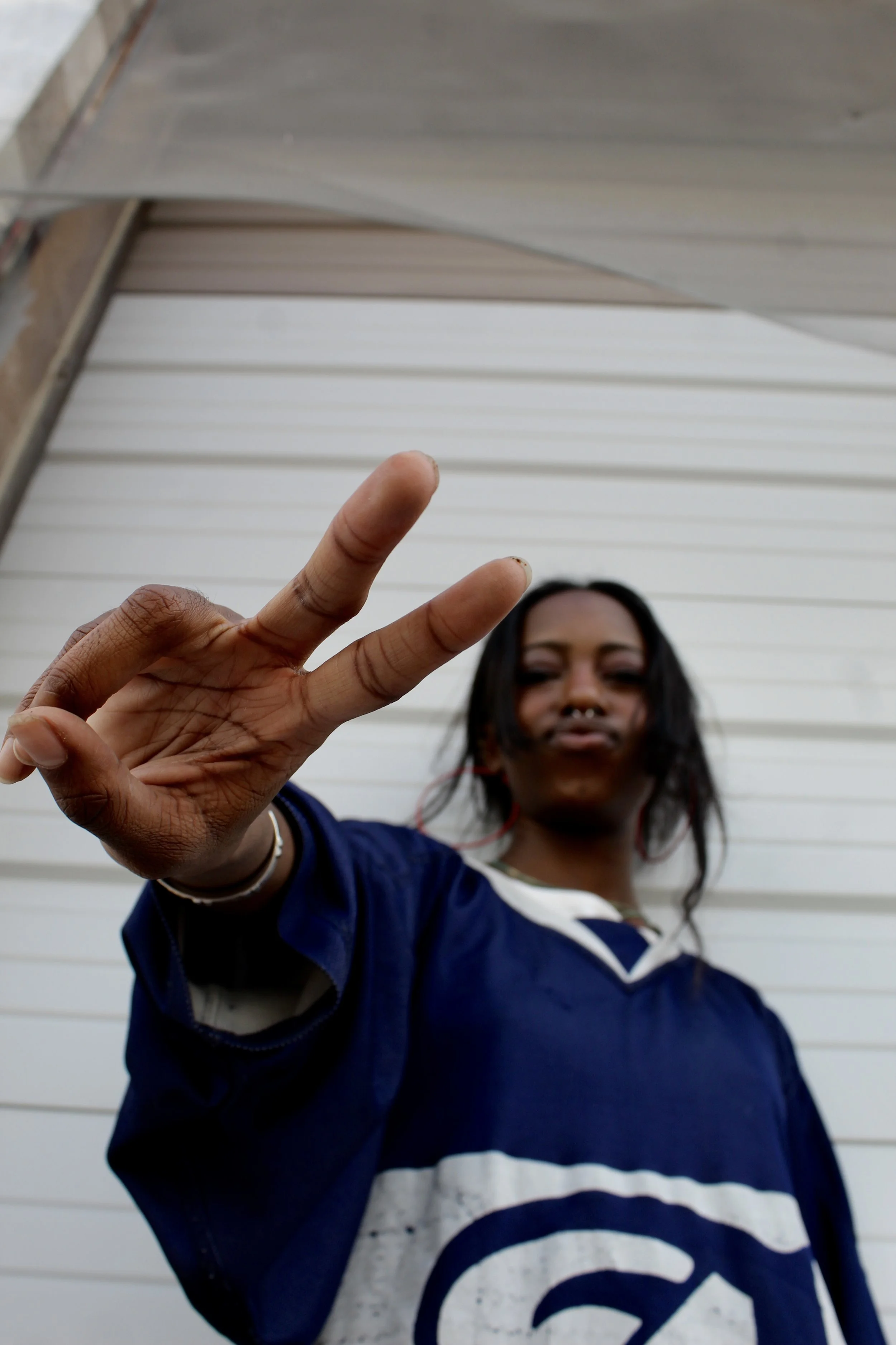 A woman with dark hair and medium skin tone making a peace sign with her hand in front of her face, standing in front of a white paneled wall.