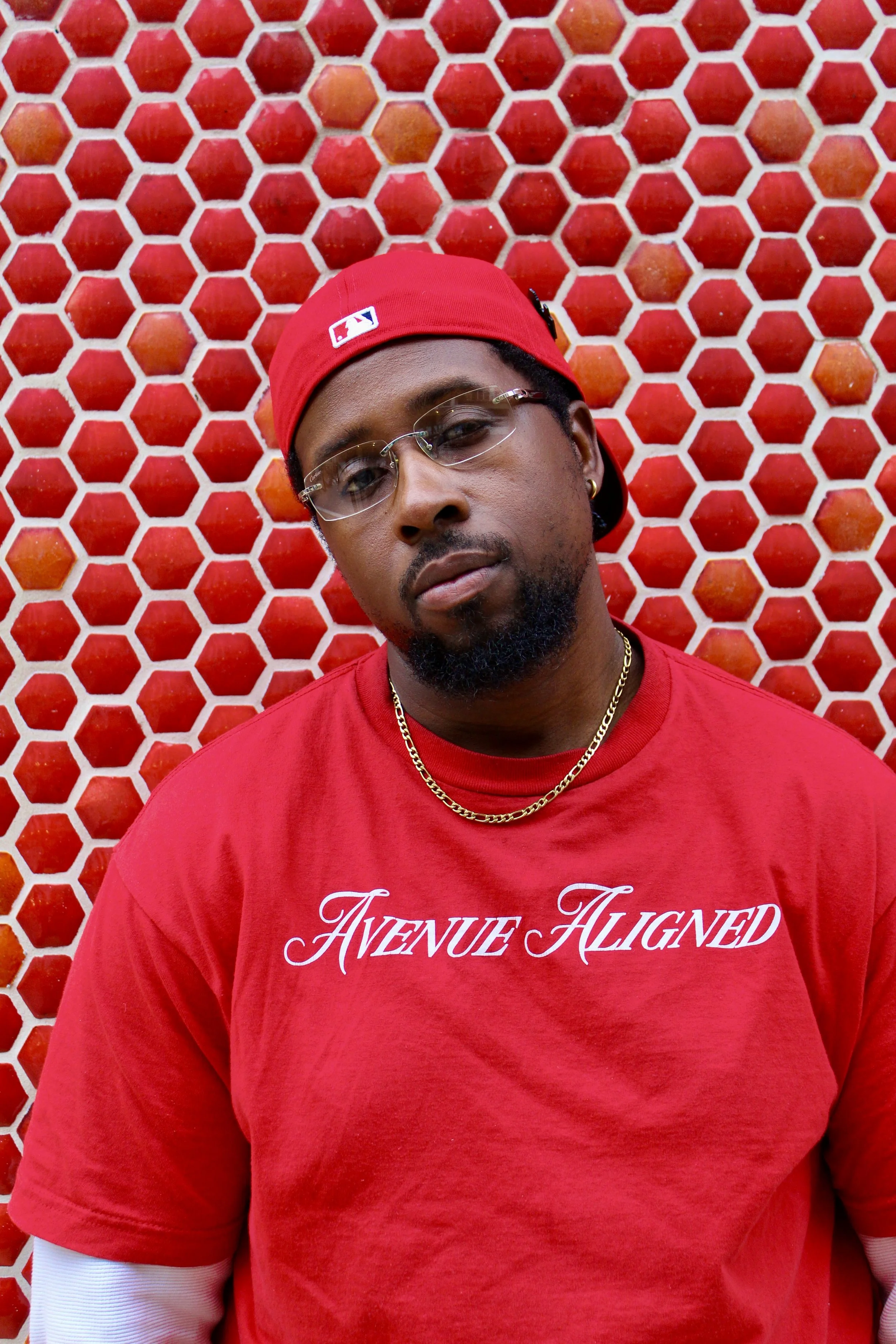 Close-up of a man wearing a red baseball cap, glasses, gold chain, and red shirt with white lettering, standing in front of a red honeycomb-patterned background.