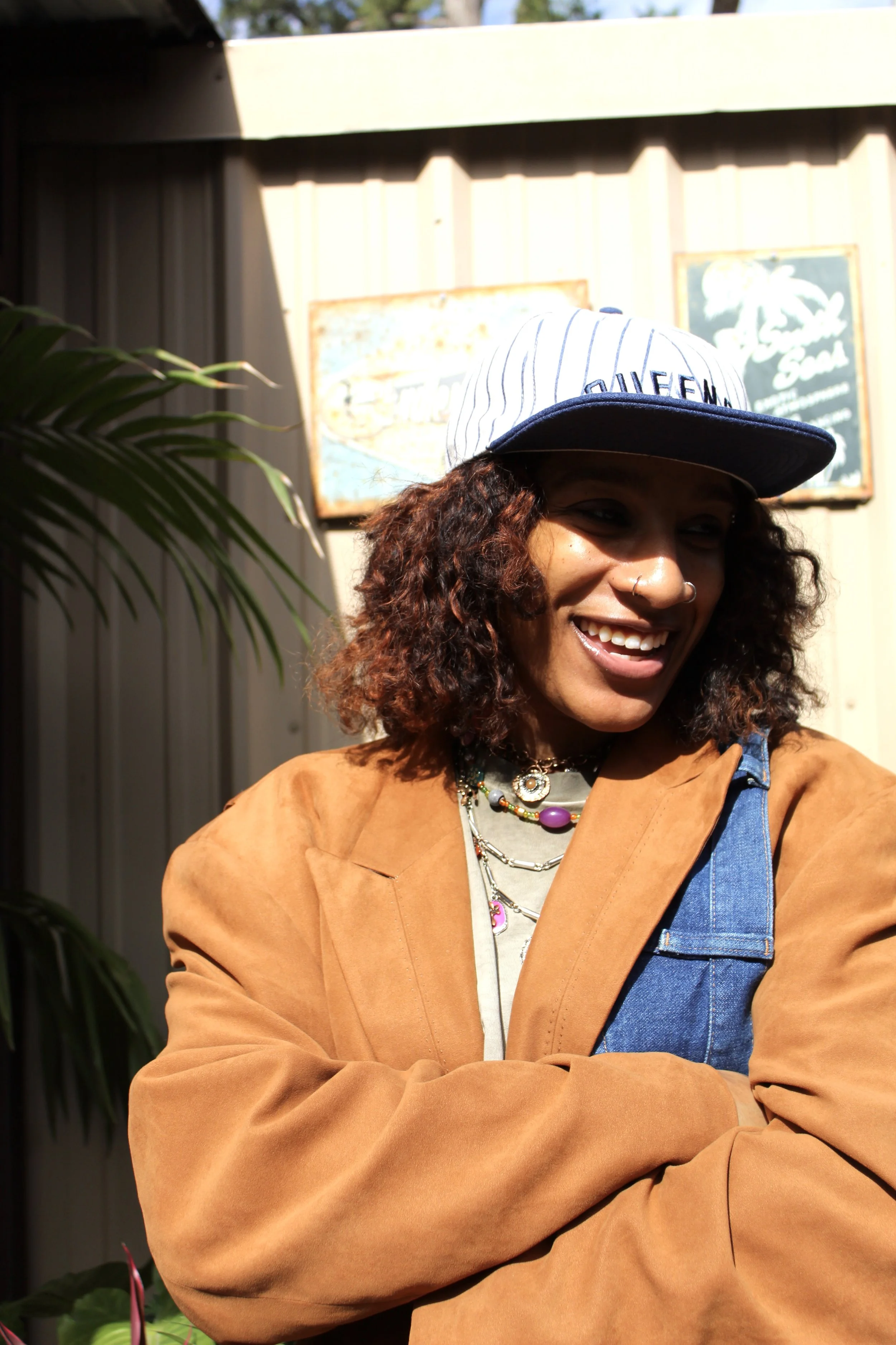 A woman with curly hair and jewelry wearing a baseball cap and a multicolored jacket, smiling outdoors with a plant and signs in the background.