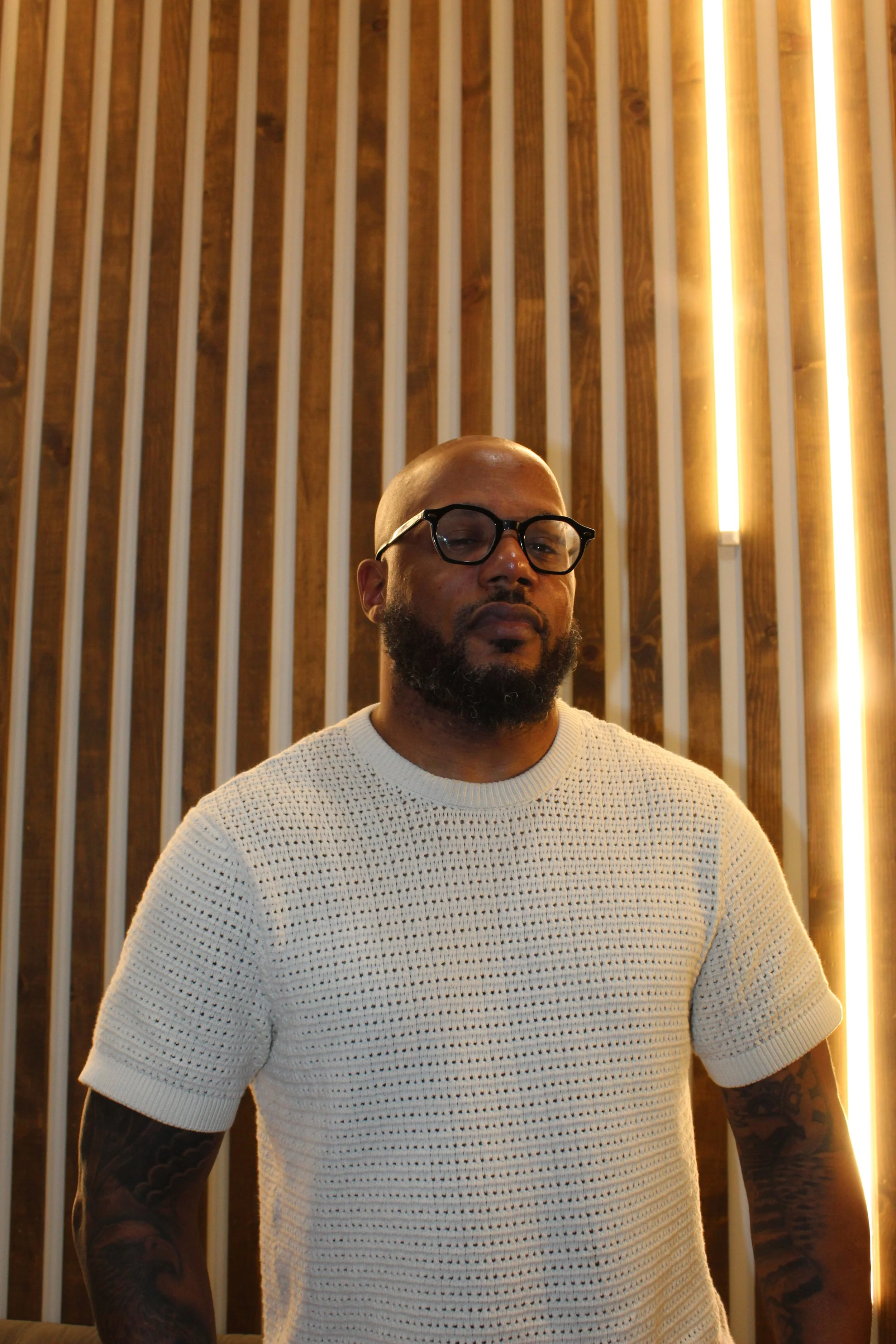 A man with a bald head, glasses, and a beard wearing a cream-colored, textured knitted short-sleeve shirt, standing in front of wooden slats with lighting accents.