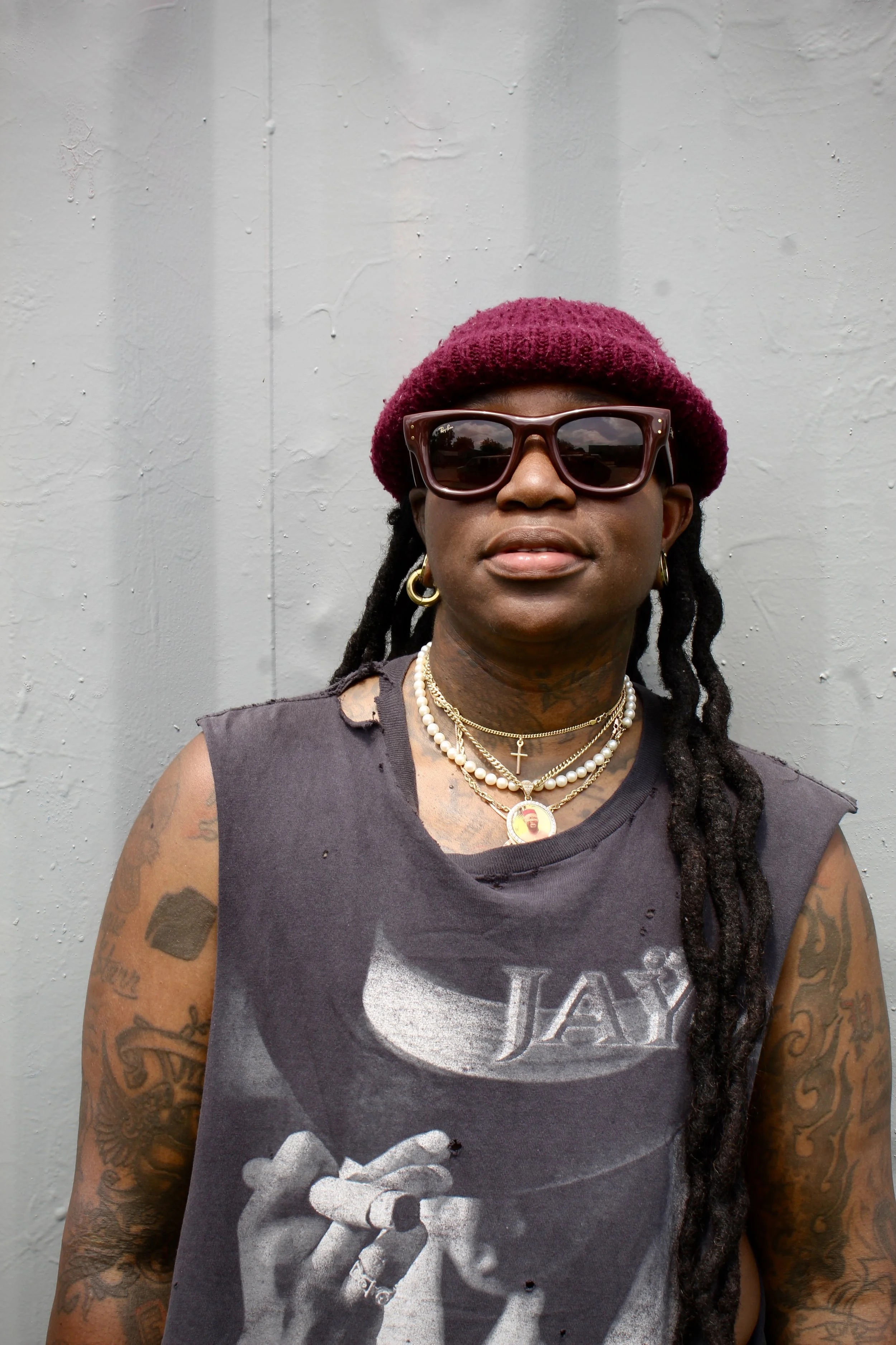 A person with dark skin wearing a purple knit beret, large sunglasses, and layered gold necklaces, standing in front of a gray wall. They are dressed in a sleeveless black shirt with a graphic print and have tattoos on their arms and neck, with long dreadlocks.