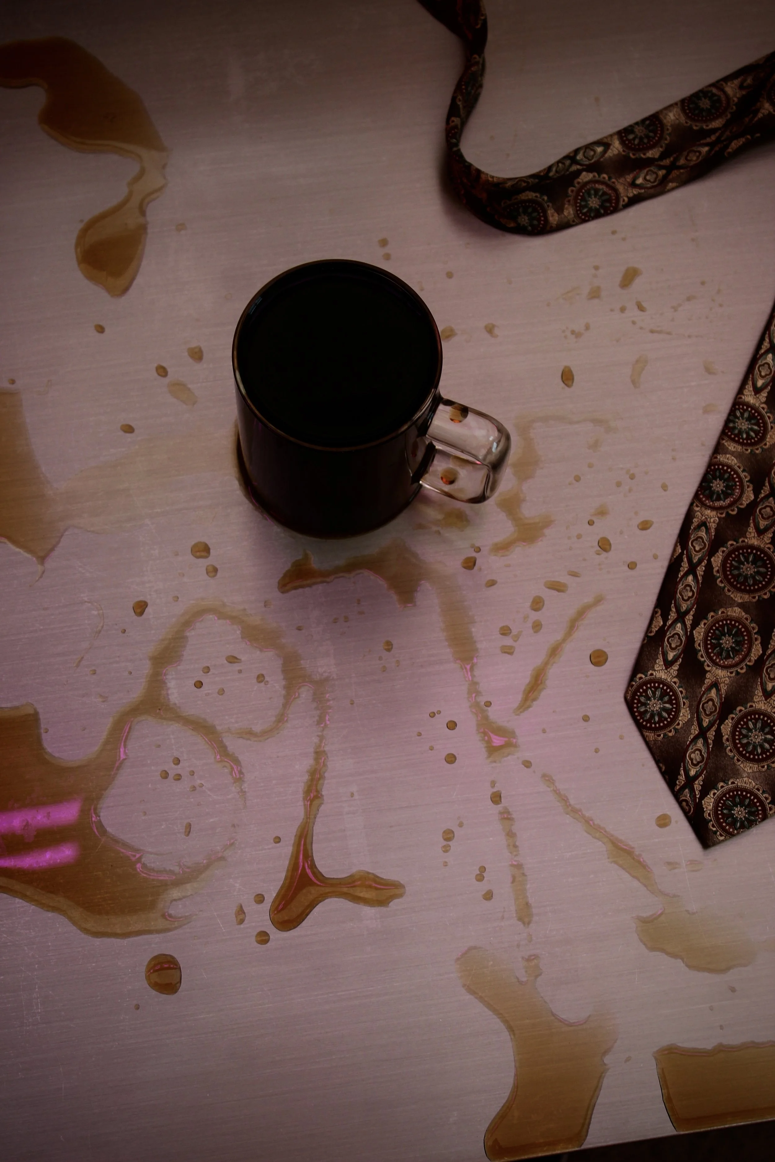 A spilled dark liquid, possibly coffee, on a wooden surface, with a coffee mug and a patterned fabric cloth nearby.