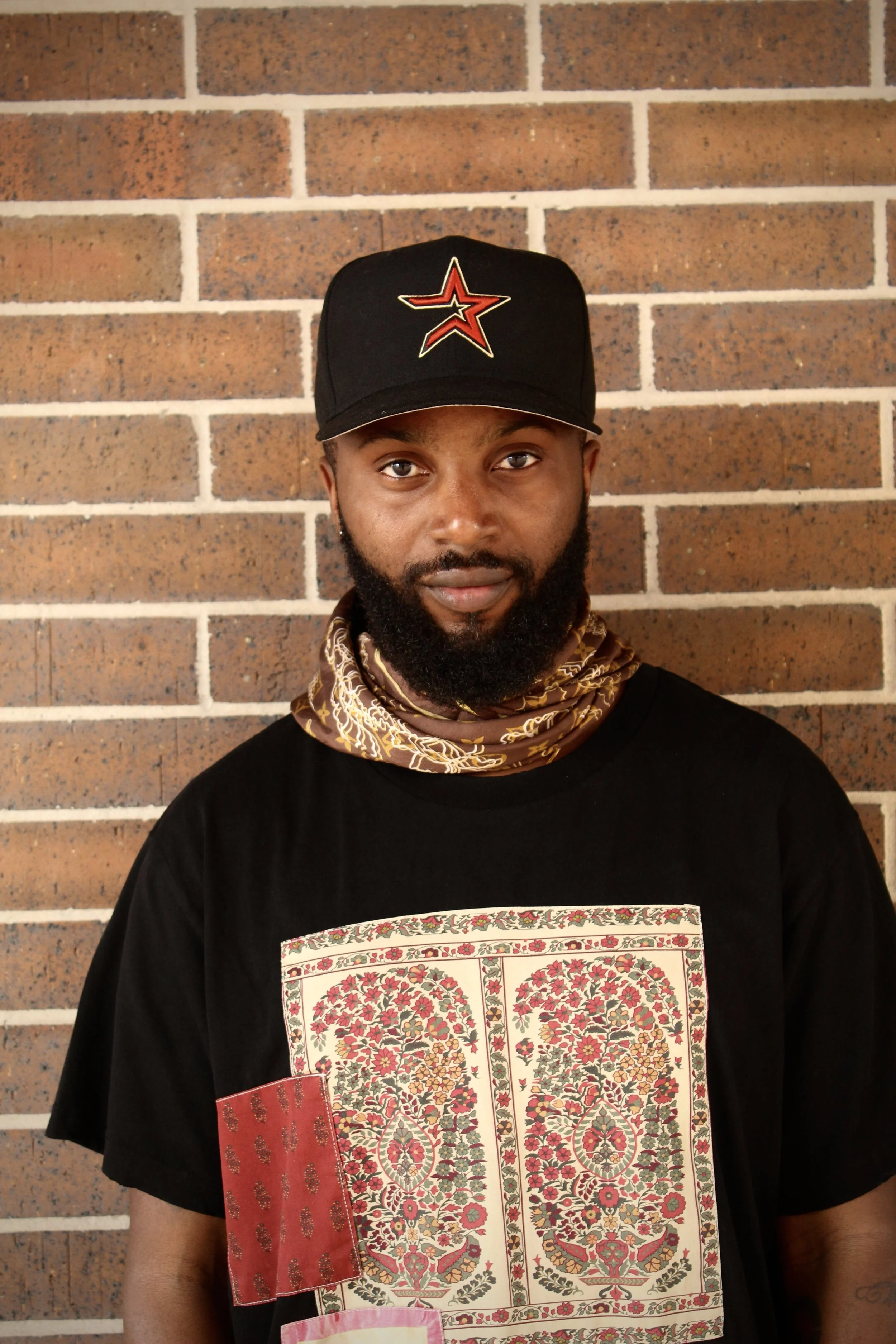 A man with a beard and mustache wearing a black cap with a red star logo, a patterned silk scarf, and a black T-shirt with a colorful floral and geometric pattern on the front, standing against a brick wall.