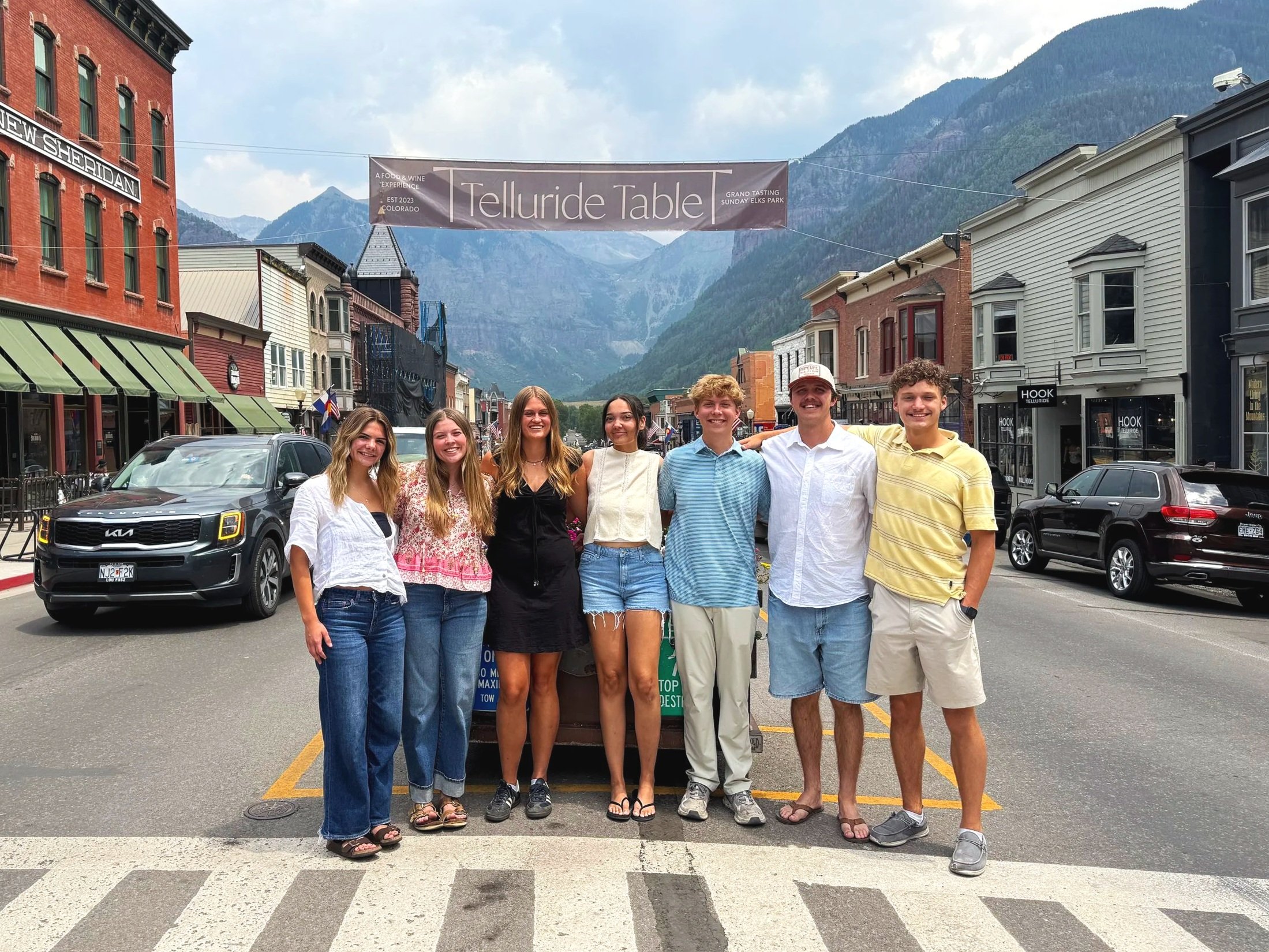 Group of seven young adults standing together on a street in a mountain town, smiling at the camera. Behind them are parked cars, storefronts, a banner overhead reading 'Telluride Table' in Grand Tasting Sunday Elks Park, and mountainous landscape in the background.