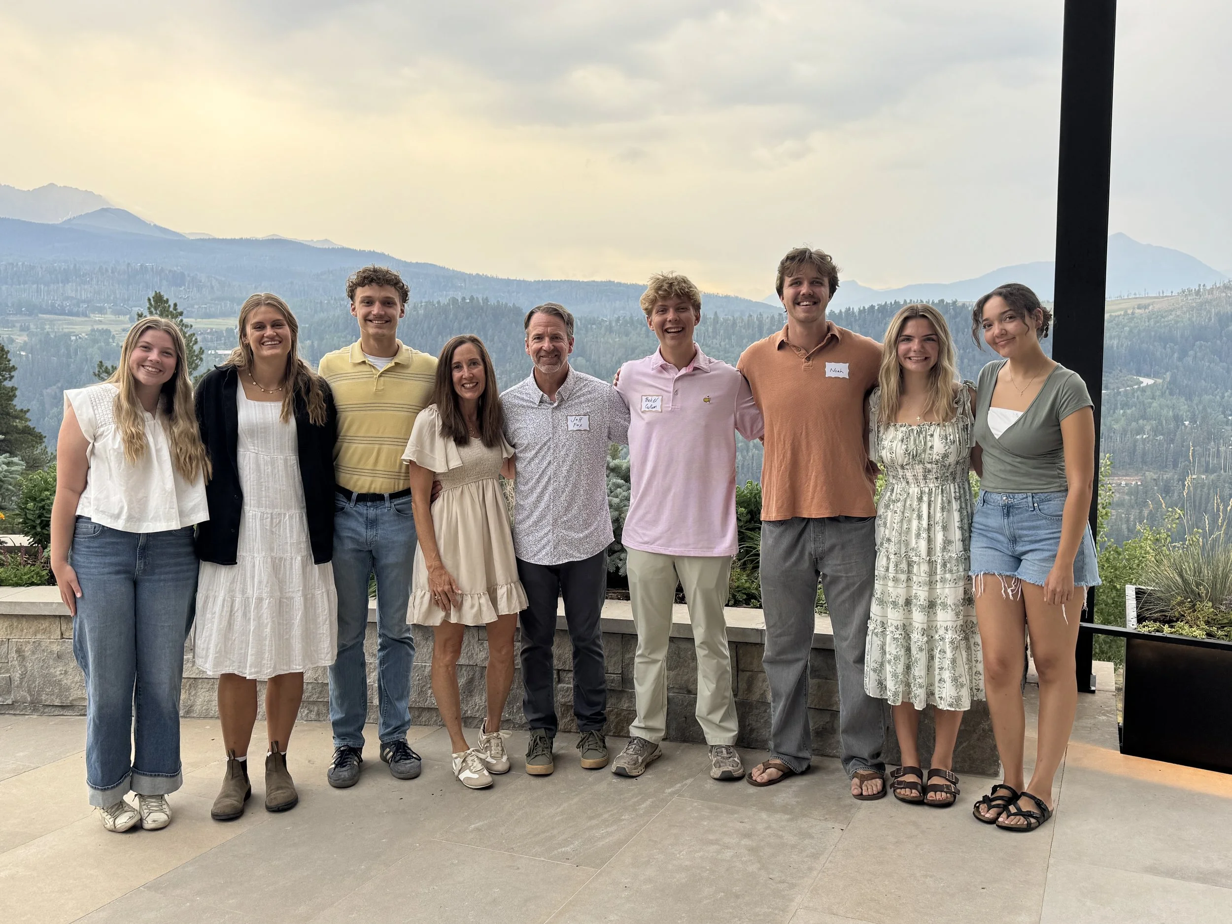 Group of nine people standing outdoors with mountains in the background, smiling and posing for a photo.