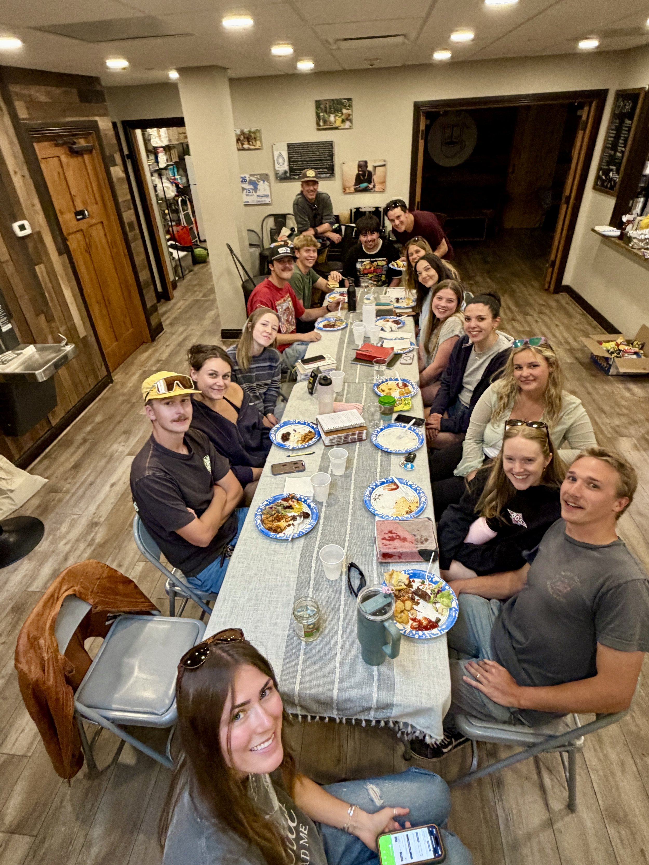 A group of young people sitting at a long dining table, smiling and enjoying a meal together in a cozy restaurant or cafe with wood accents and artwork on the walls.