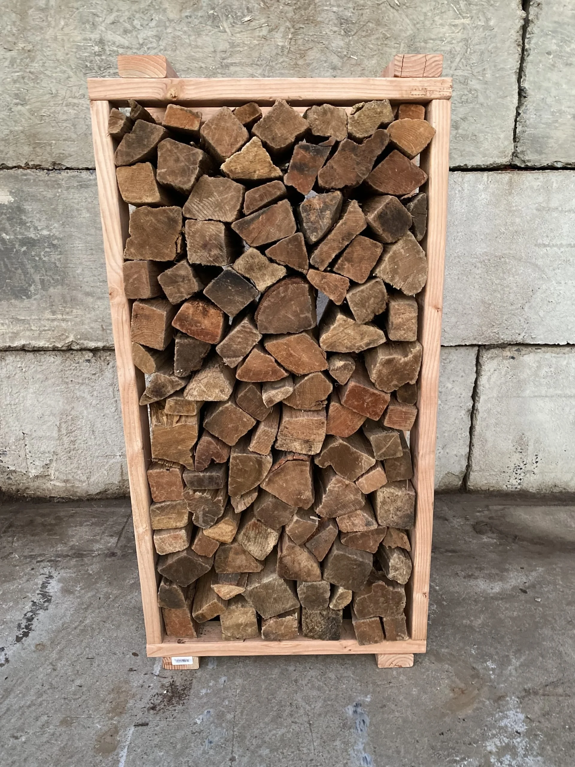 Stack of chopped firewood inside a wooden frame, placed on a concrete floor against a stone wall background.
