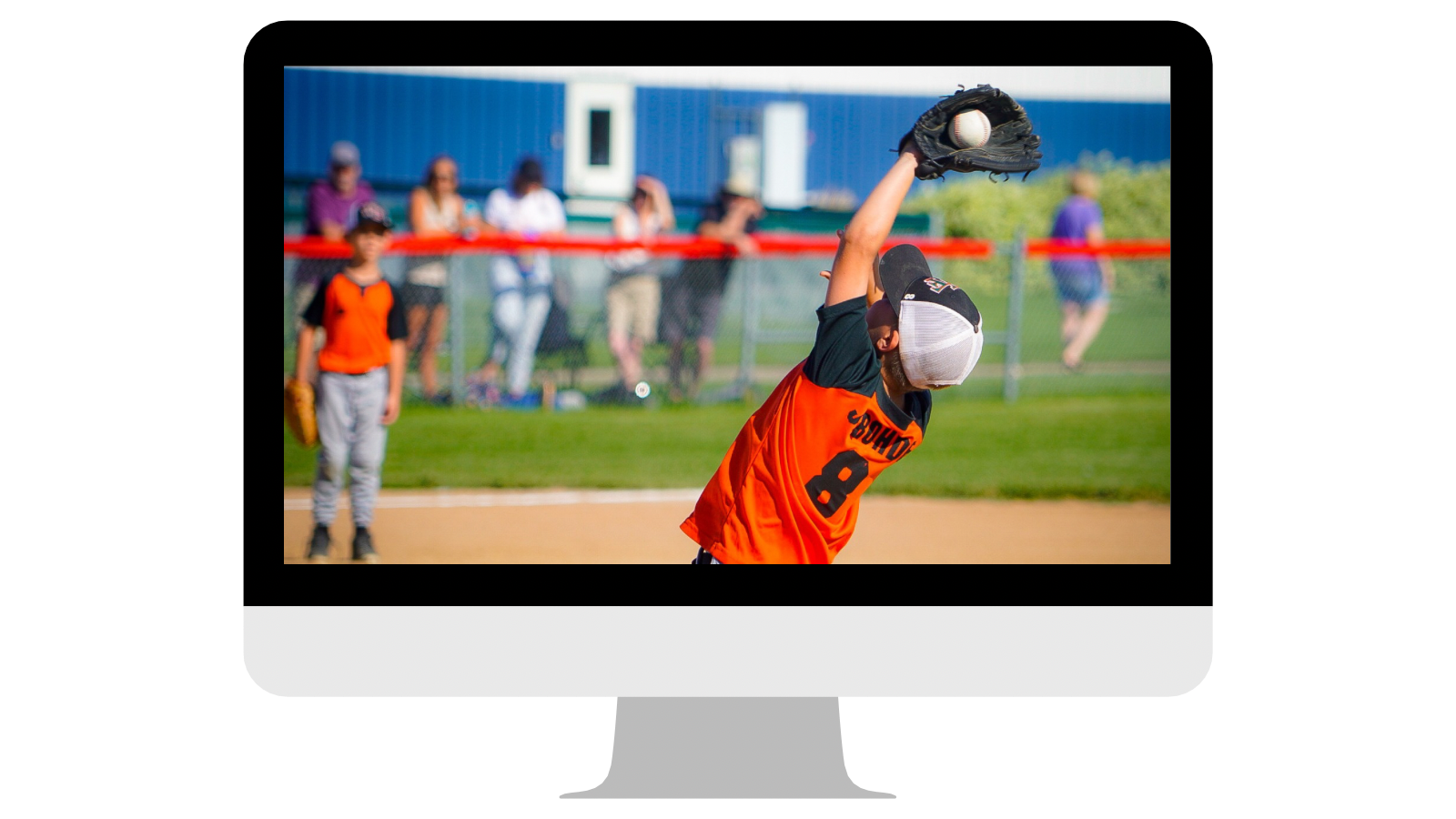 A young baseball player in an orange jersey with the number 8 on it, reaching up with a glove to catch a baseball on a softball field during a game. Spectators are blurred in the background behind a chain-link fence.