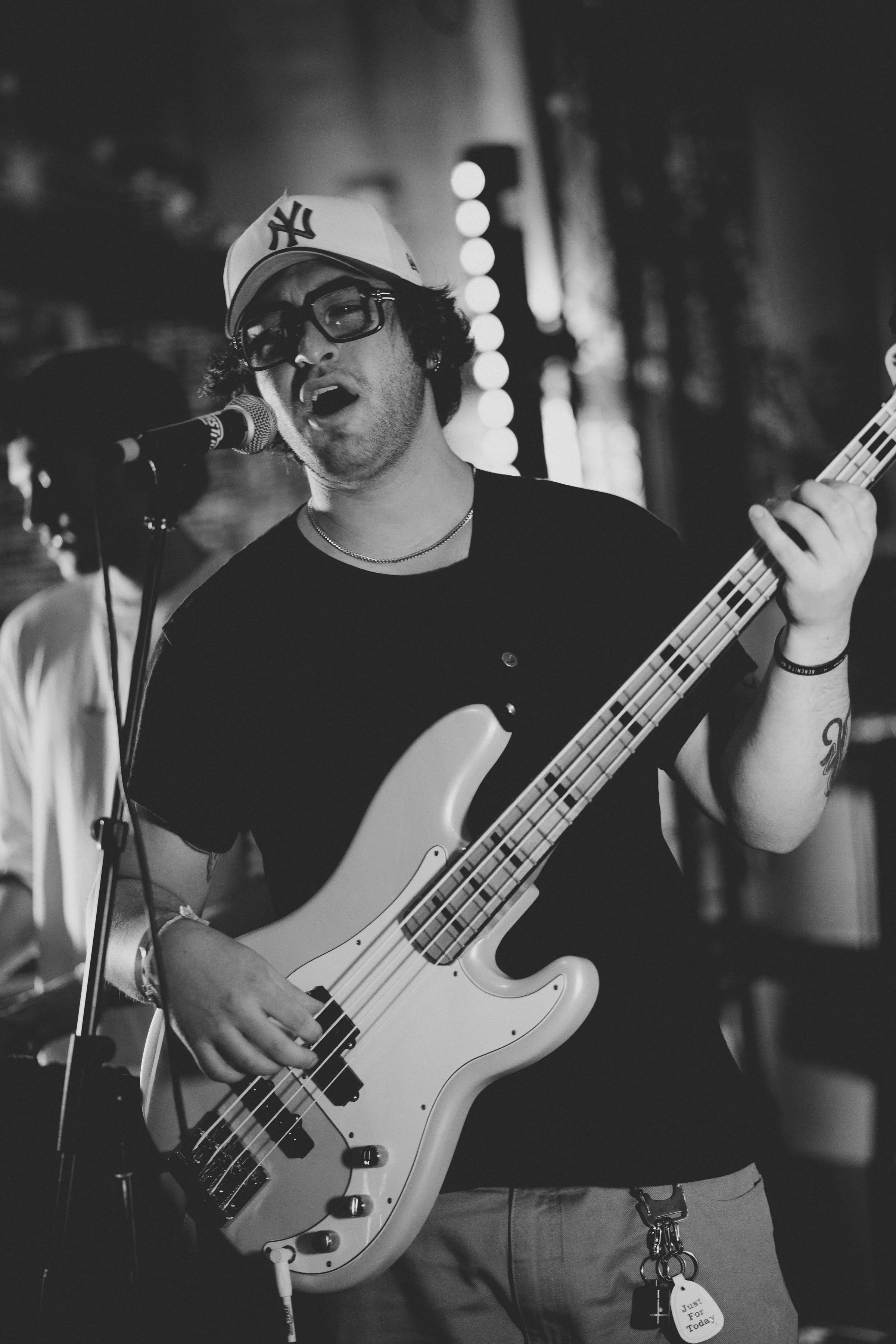 A man singing and playing a bass guitar on stage, wearing a white New York Yankees cap and glasses, with background lights and a blurry person nearby.