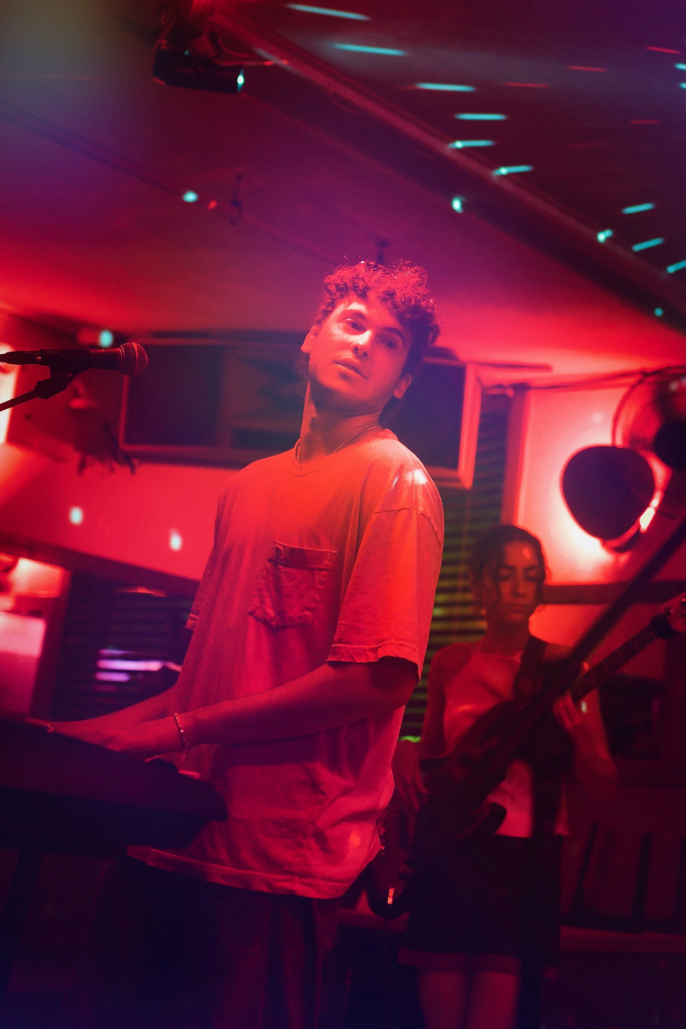 A young man playing keyboard and a woman playing bass guitar in a dimly lit room with red and blue neon lights, creating a vibrant atmosphered.