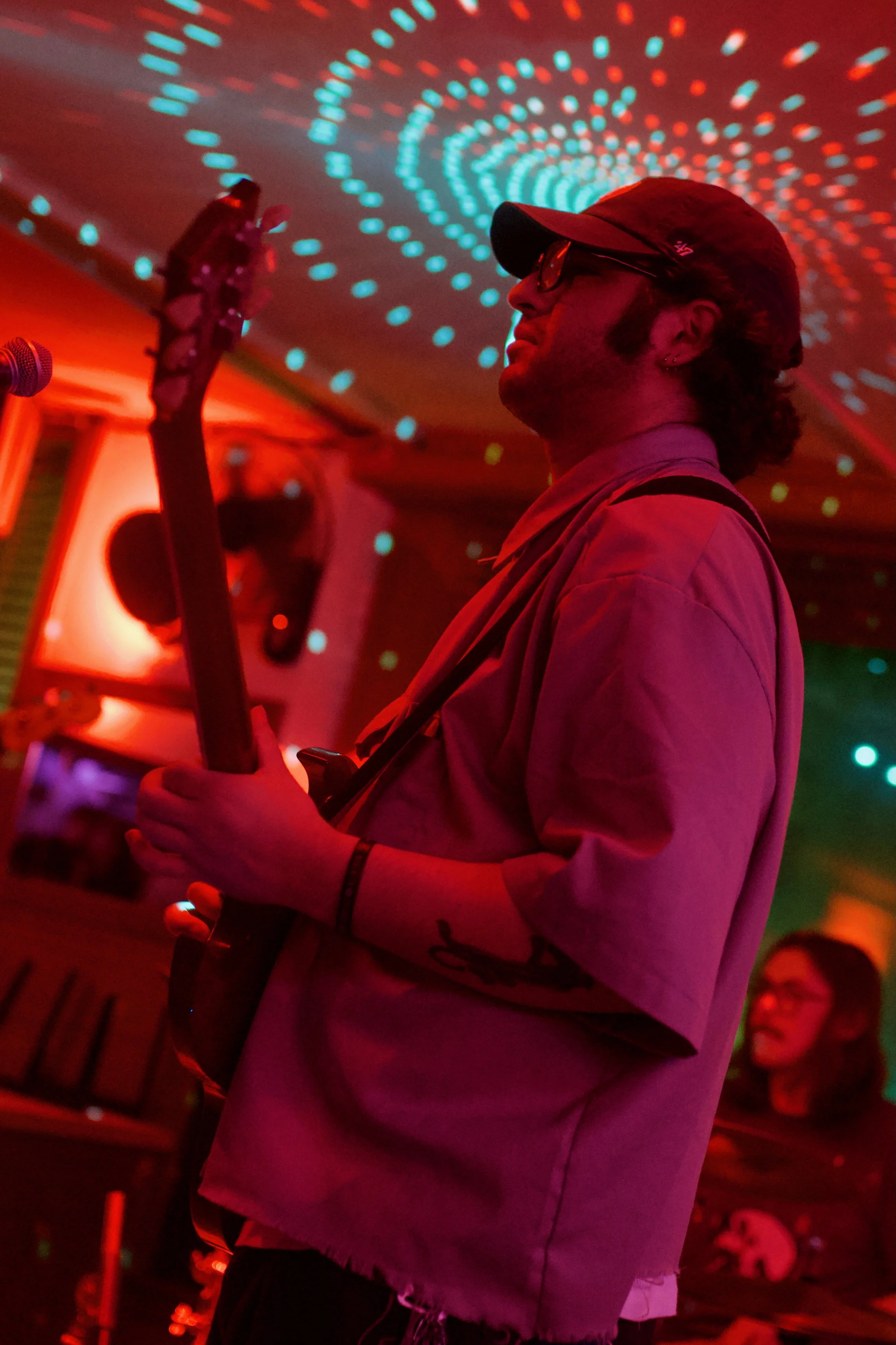 A man wearing a cap and glasses playing an electric guitar in a dimly lit room with colorful, swirling LED lights on the ceiling, and a woman in the background.
