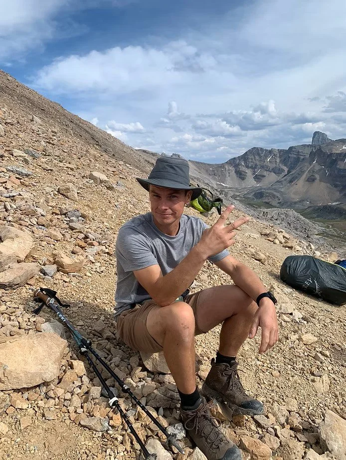 A person in hiking gear, including a wide-brimmed hat, gray t-shirt, shorts, and hiking boots, sitting on a rocky mountain terrain with a mountain landscape in the background, making a peace sign with their right hand.